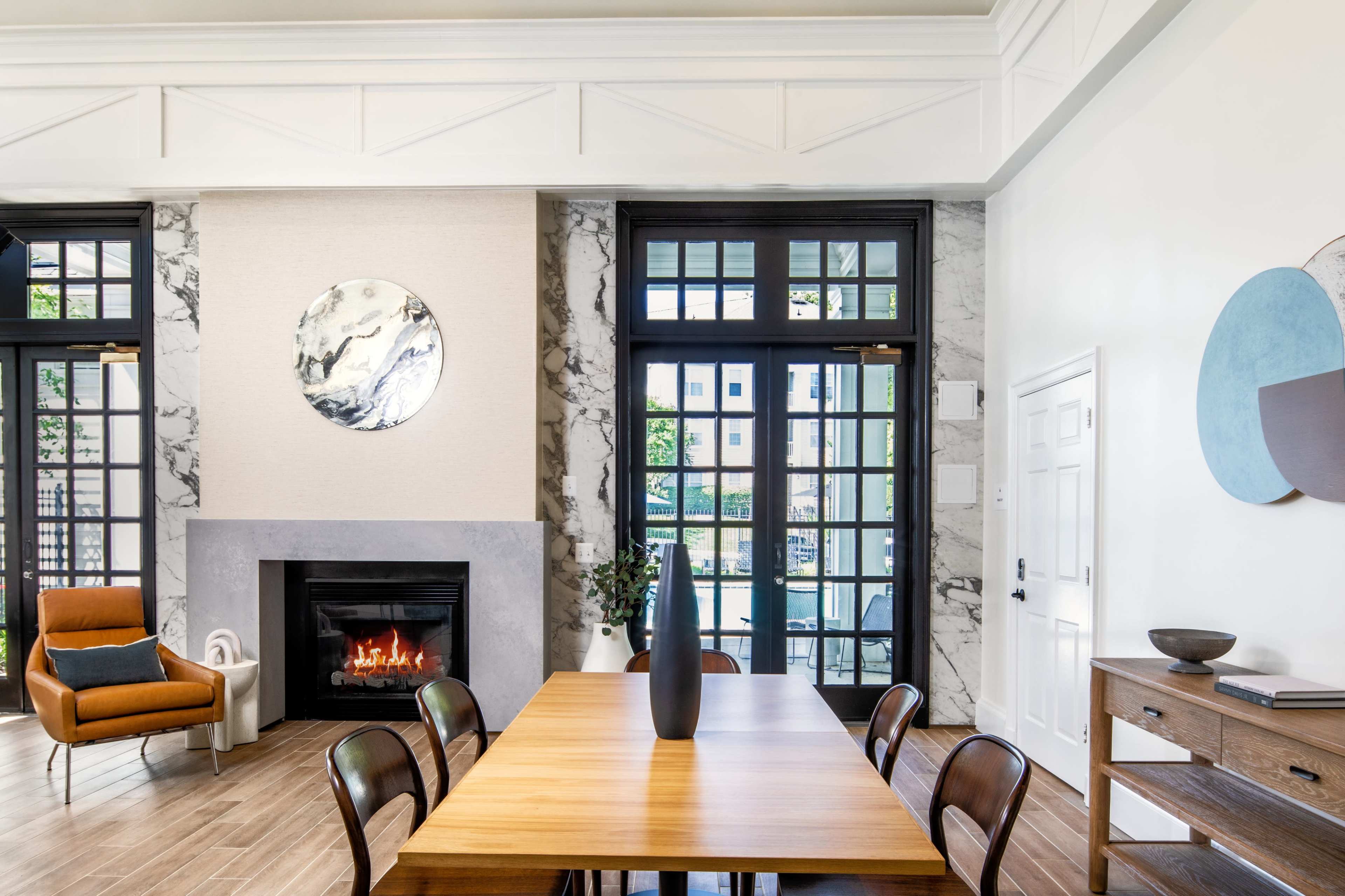 The image shows a modern interior living space featuring a table with wooden chairs, a fireplace, and large windows with a view of an outdoor area.