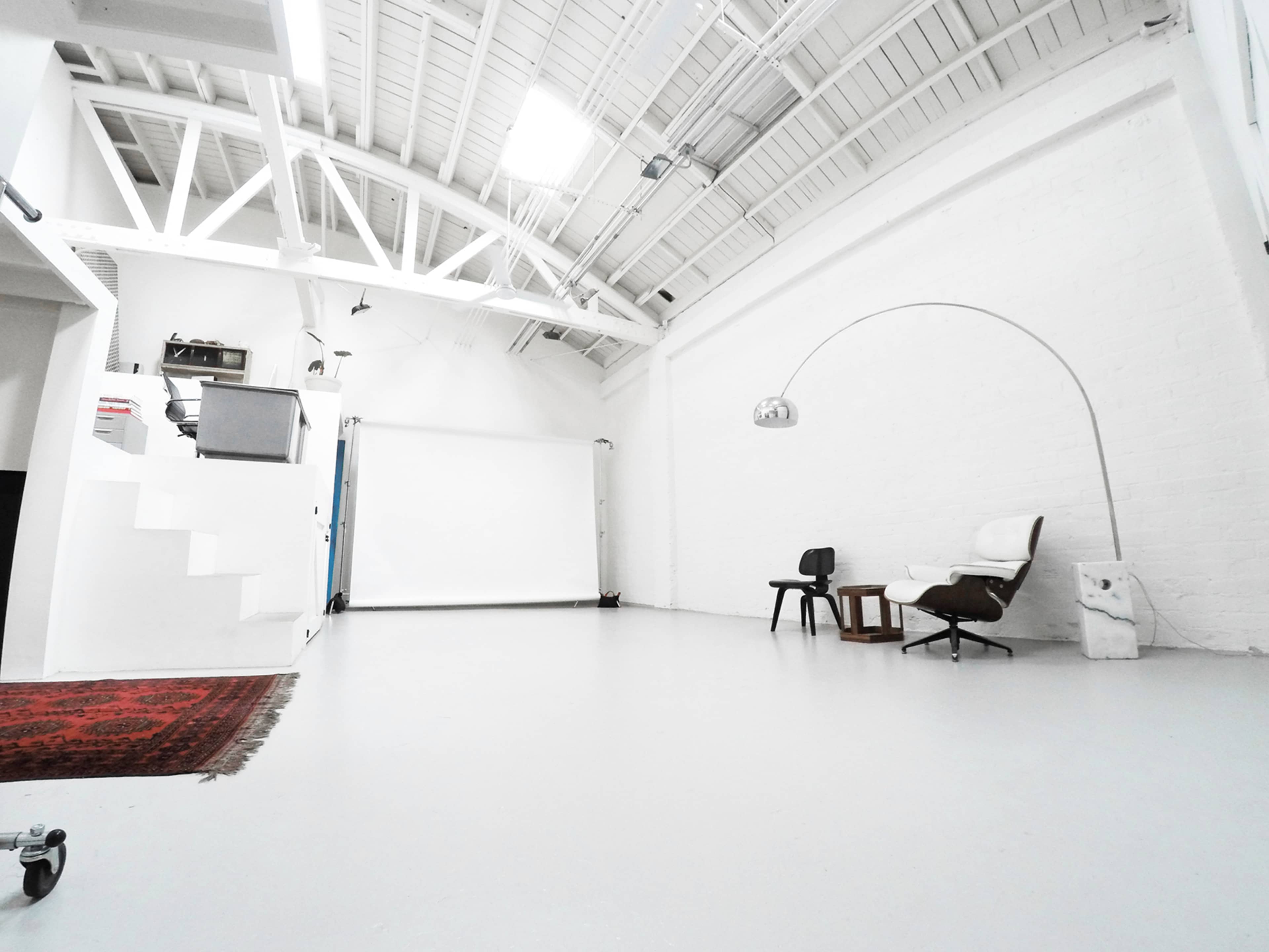 A spacious, minimalist studio with a white-painted interior, featuring a projector screen, a staircase, and two chairs.