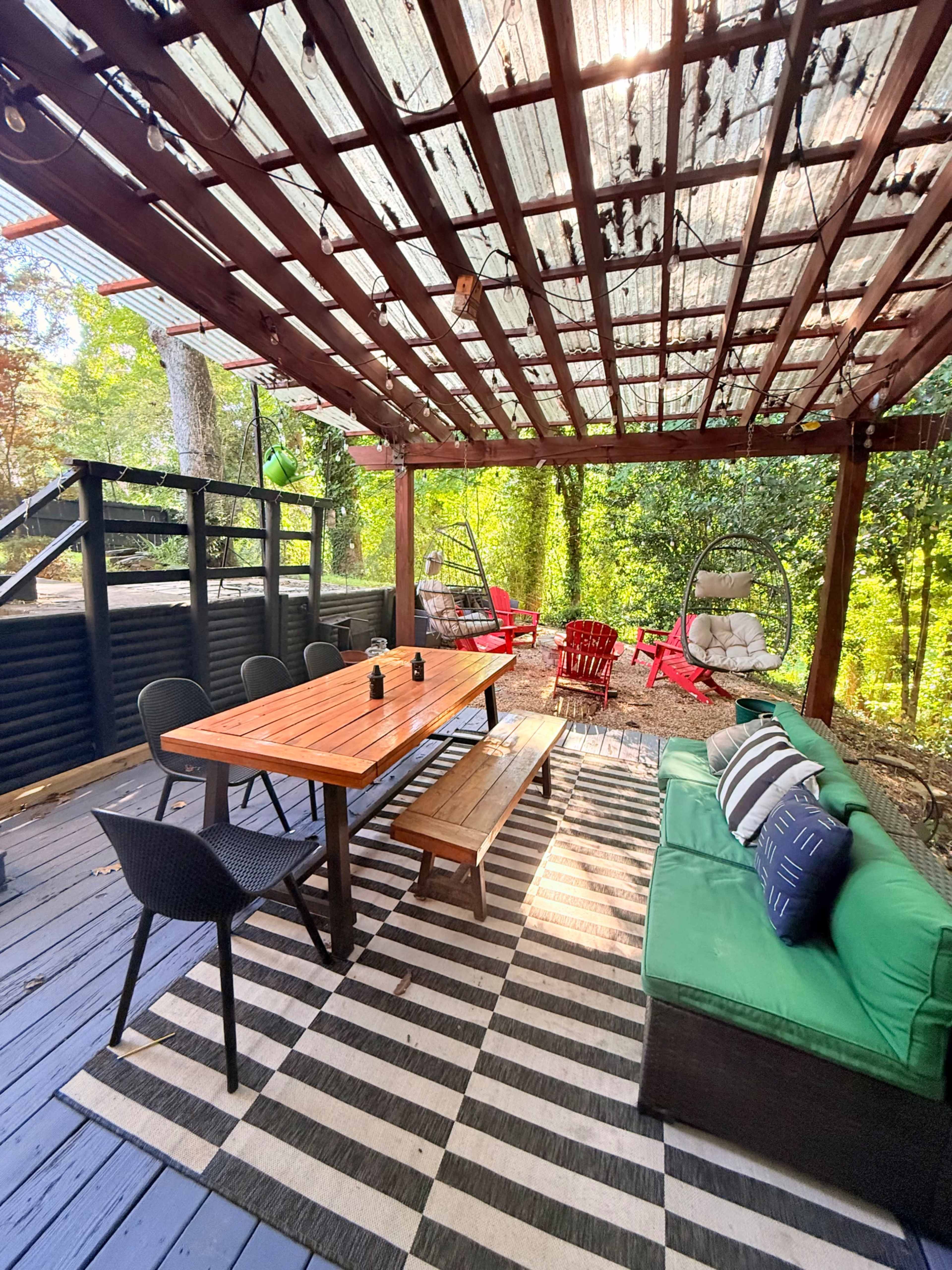A covered outdoor dining area features a wooden table and benches, surrounded by black and green seating, with hanging chairs and a striped rug.