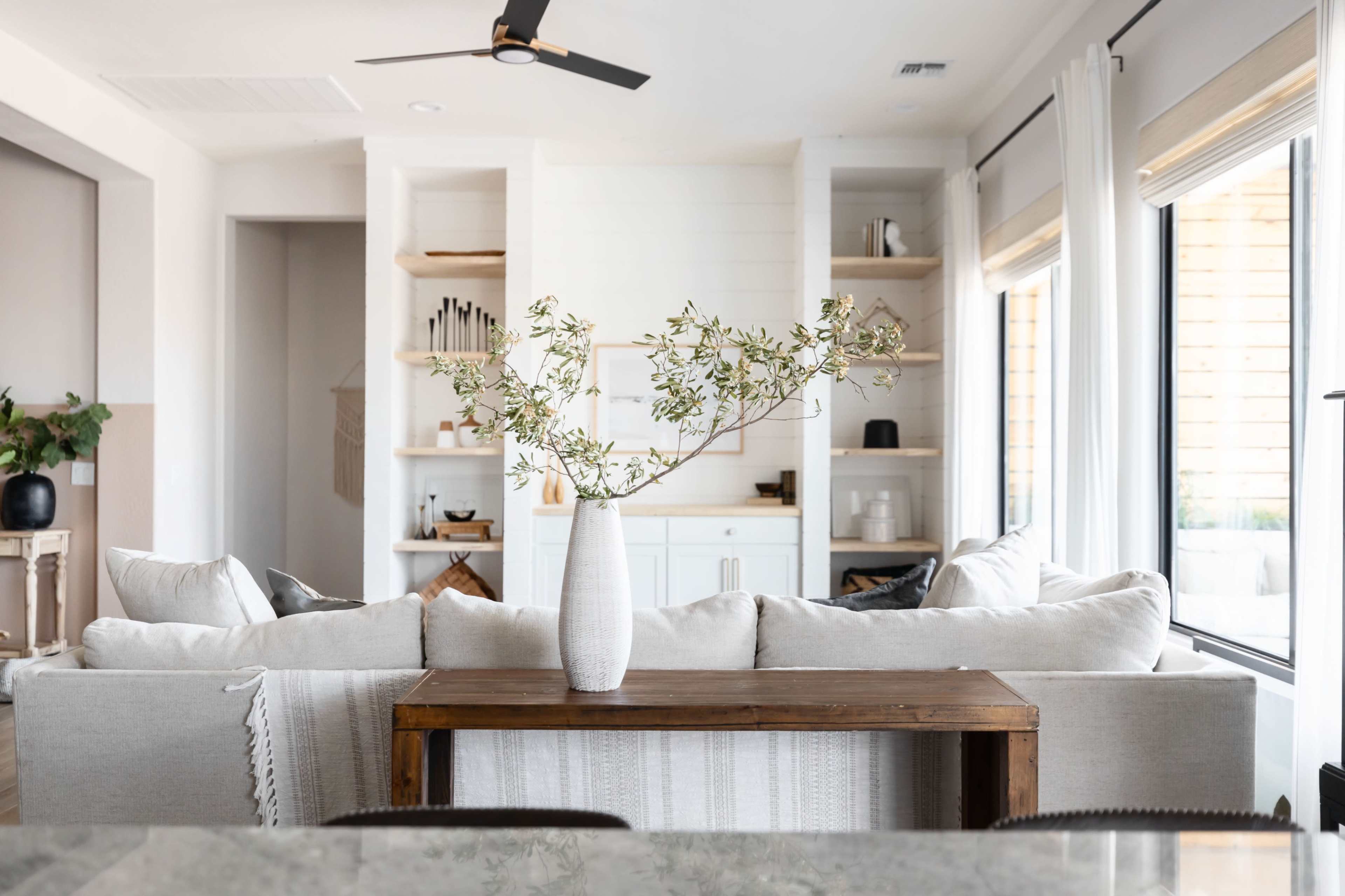A light-filled living space features a beige sofa with a wooden table in front, adorned with a vase of greenery, and a backdrop of open shelves and windows.