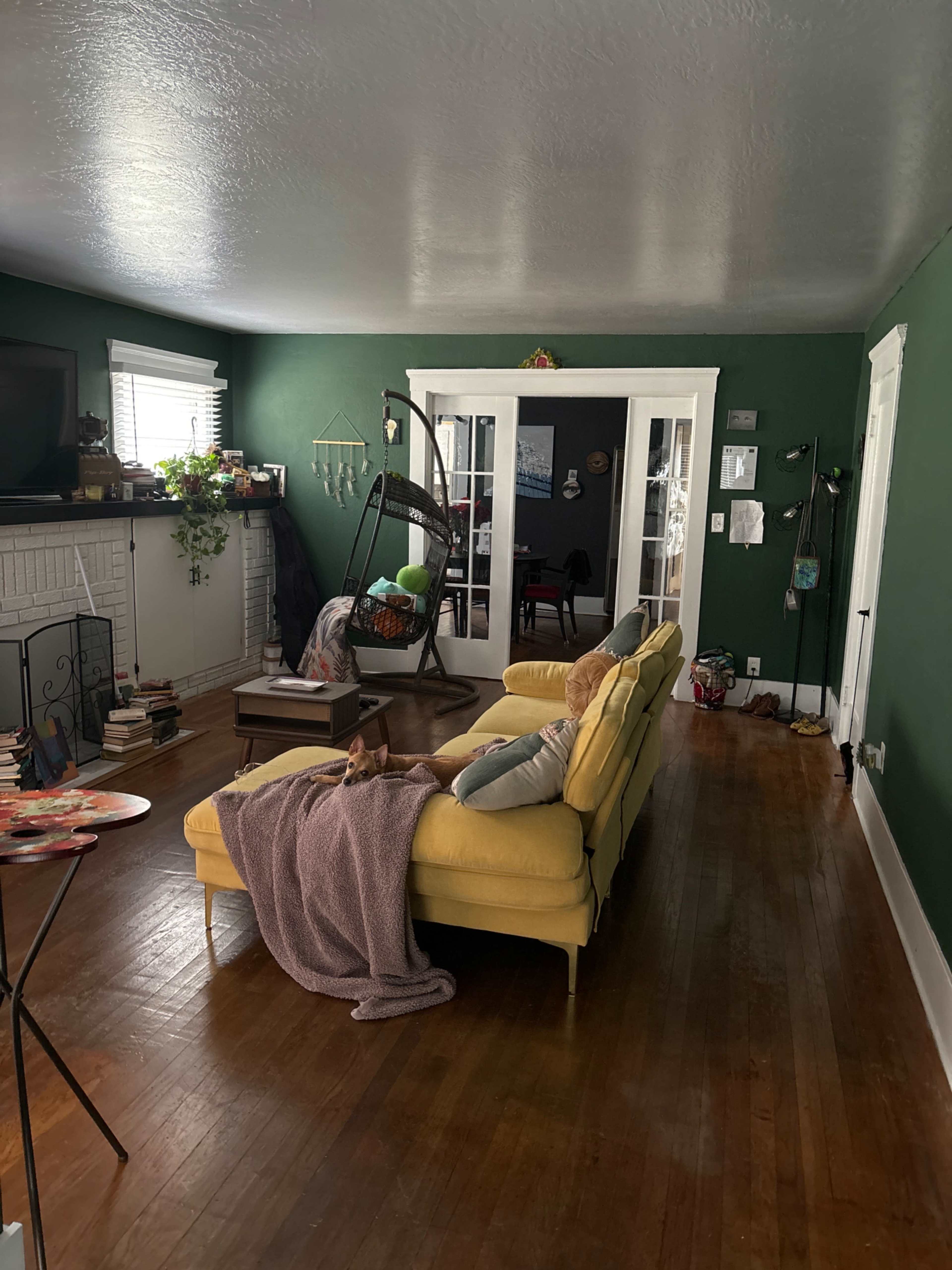 A cozy living room with a yellow sofa, a green accent wall, and wooden flooring, featuring a television and various decorative items.