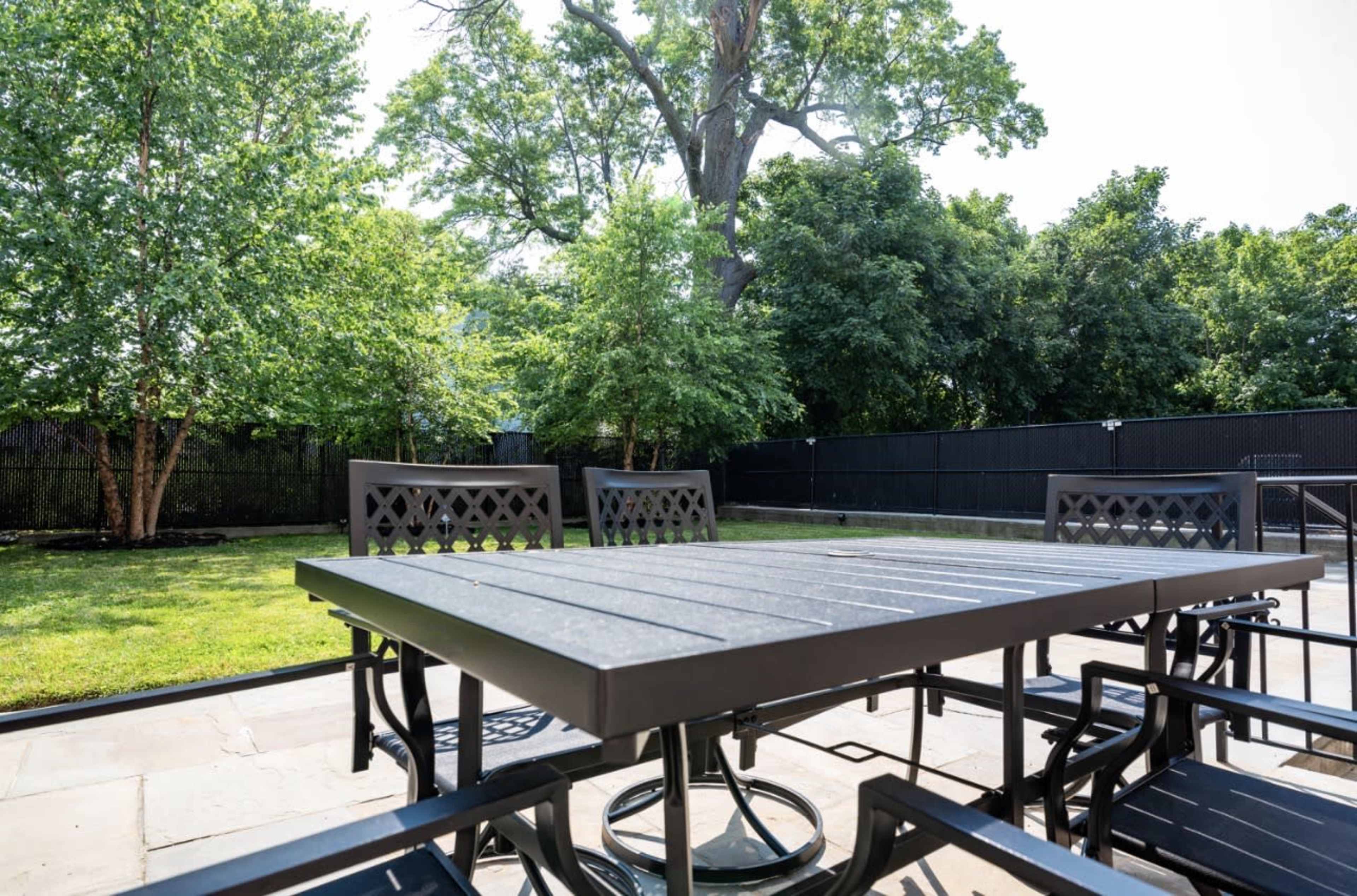 A metal patio table surrounded by chairs is set on a stone surface with a grassy backyard and trees in the background.