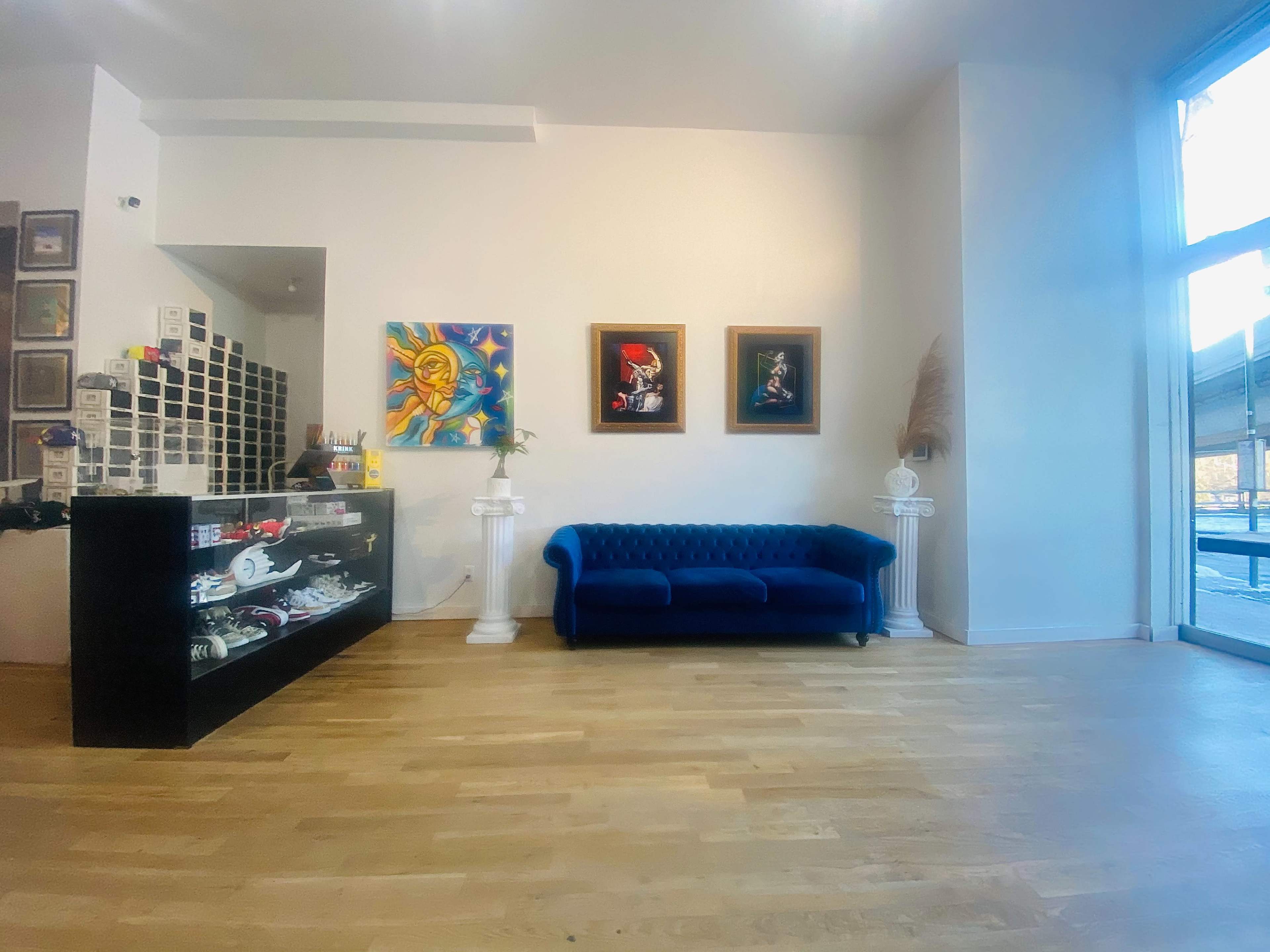 A modern interior space featuring a blue sofa, two framed artworks, and a glass display counter, all set against a light-colored wall and wooden floor.