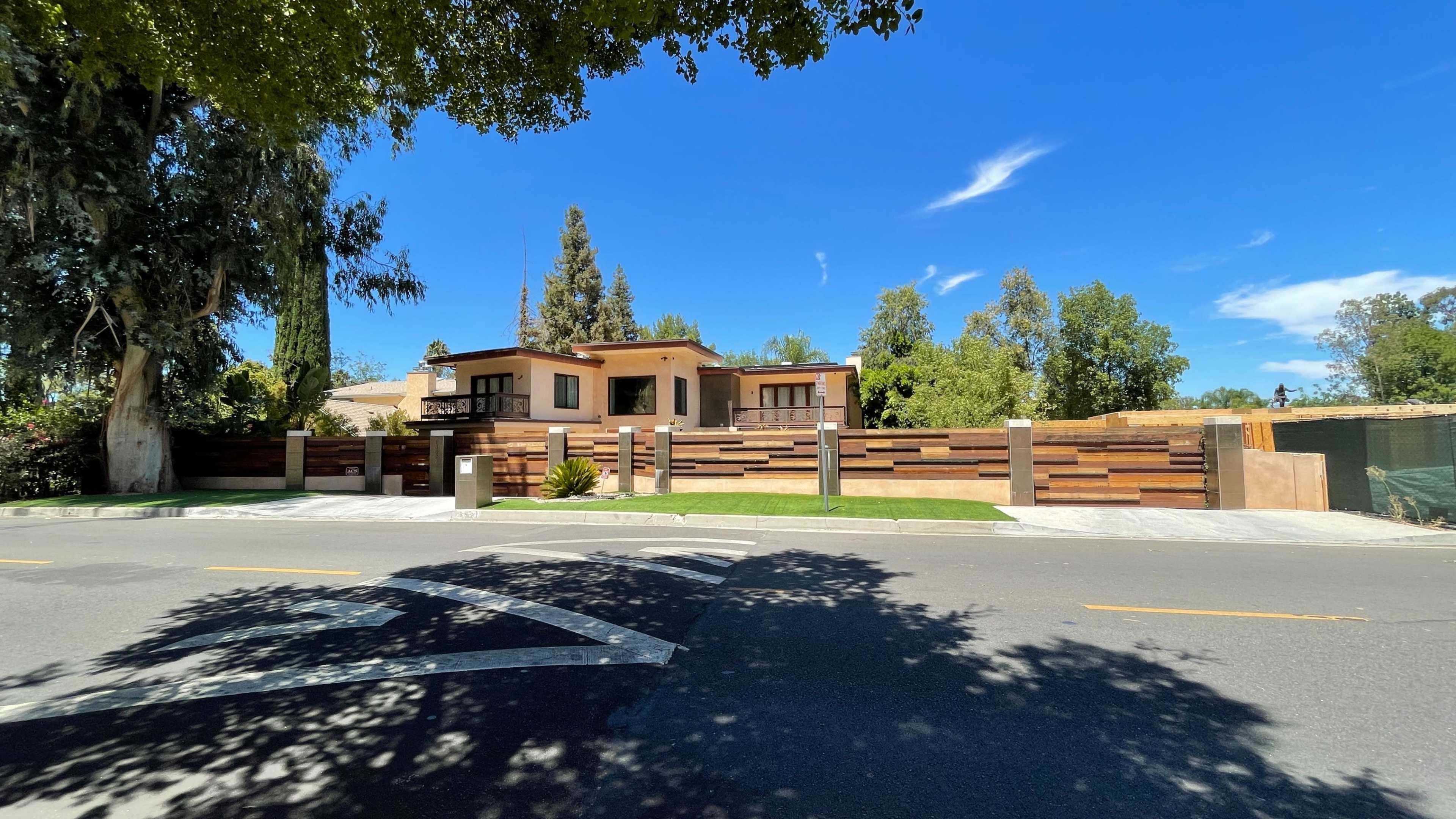 The image shows a modern house surrounded by greenery and a wooden fence, viewed from the street on a sunny day.