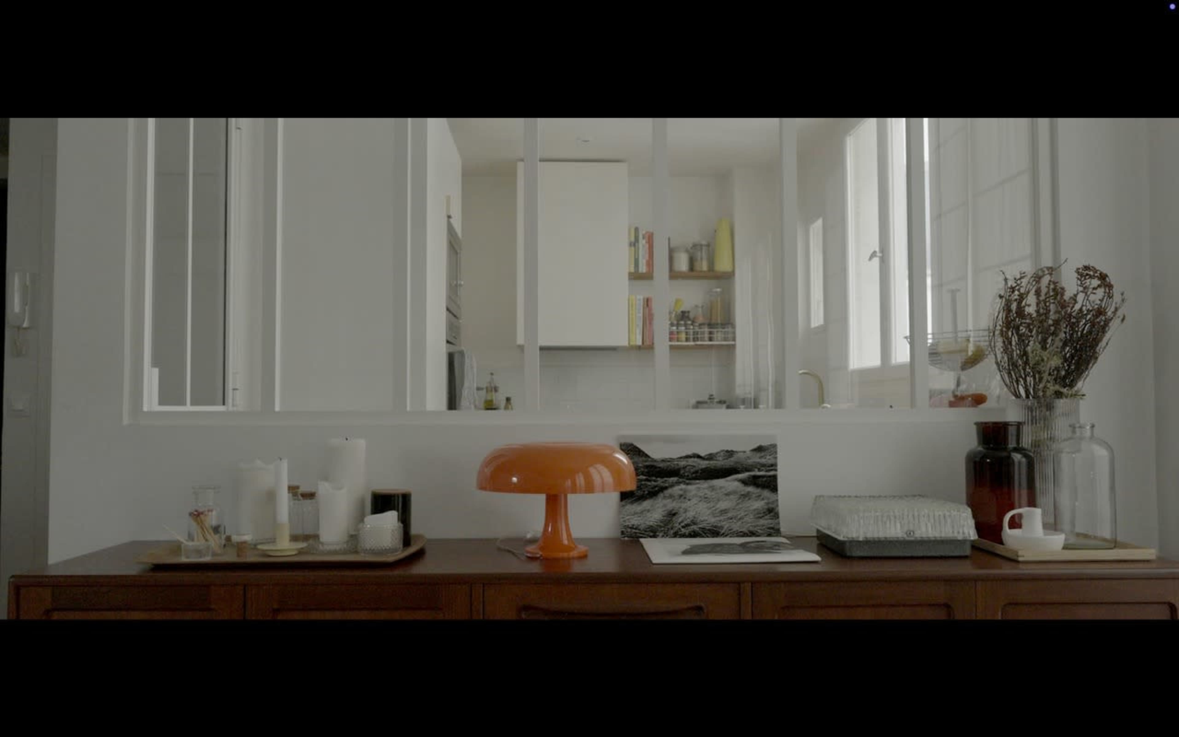 A wooden sideboard displays candles, a glass jar, and a lamp, while a kitchen is visible through a glass partition in the background.