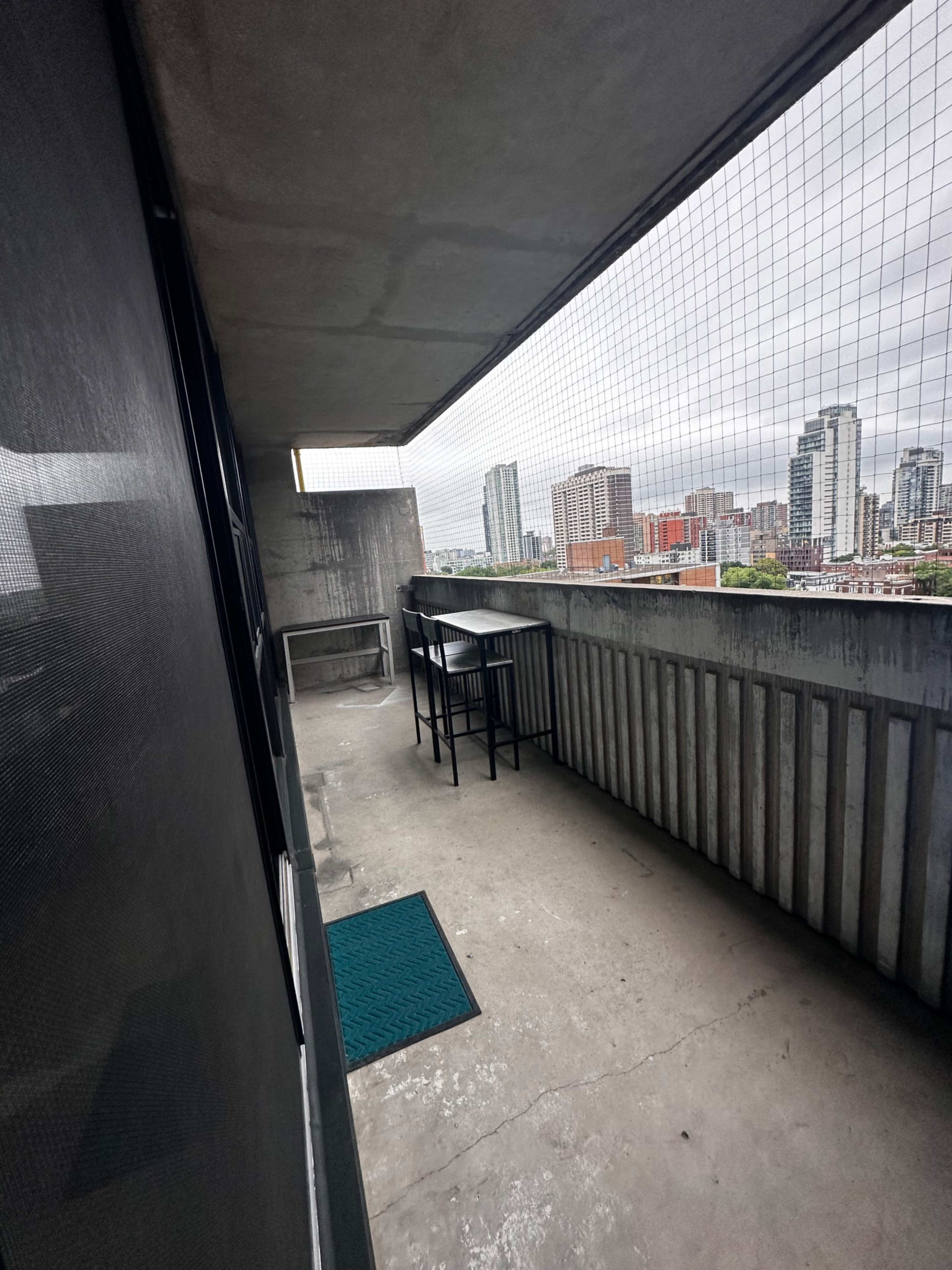 The image shows a concrete balcony with a small table and two chairs, overlooking a city skyline under a cloudy sky.