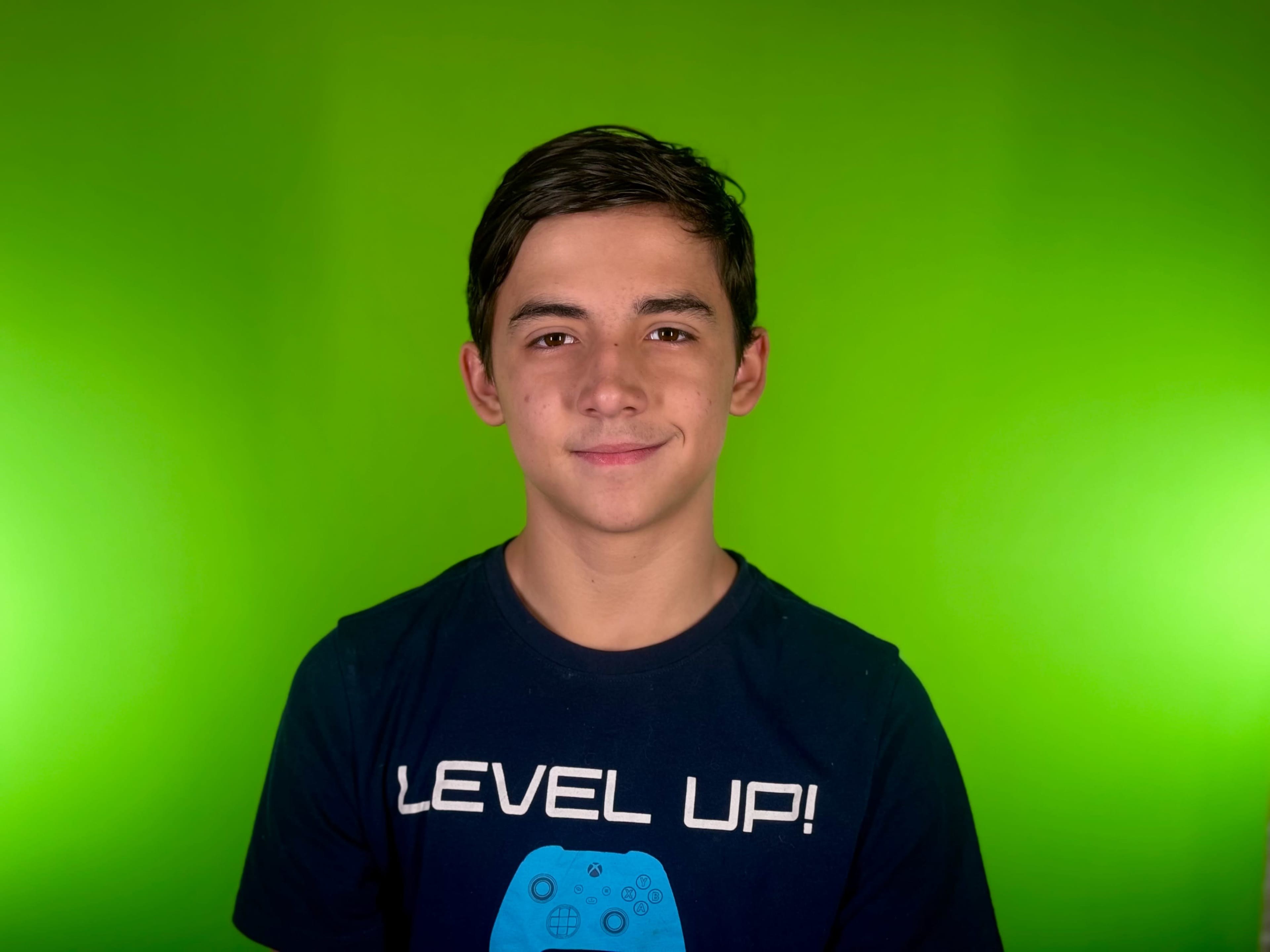 A young boy with short hair wearing a blue "LEVEL UP!" graphic t-shirt stands against a bright green background.
