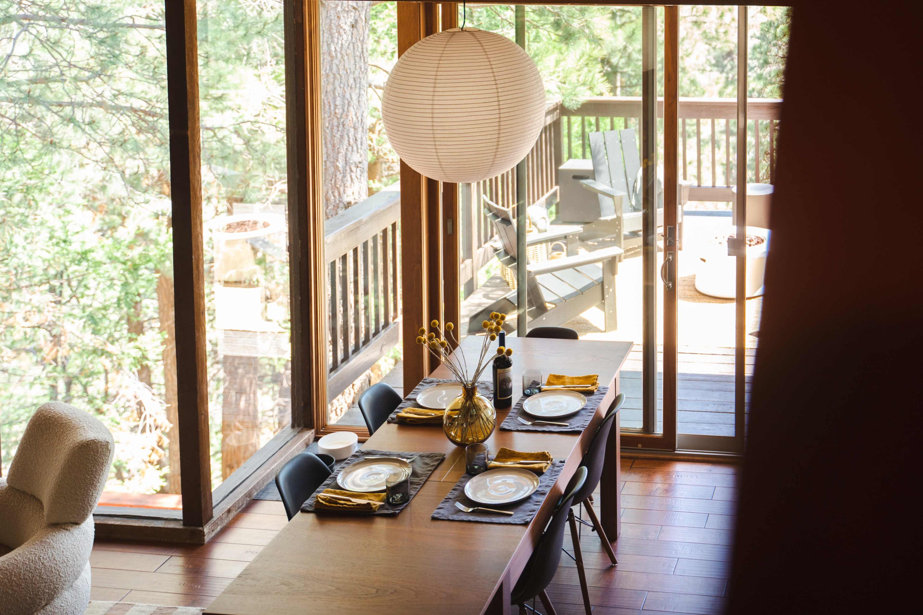 A dining table is set with plates and decorative items, next to large windows overlooking a wooden deck and outdoor scenery.