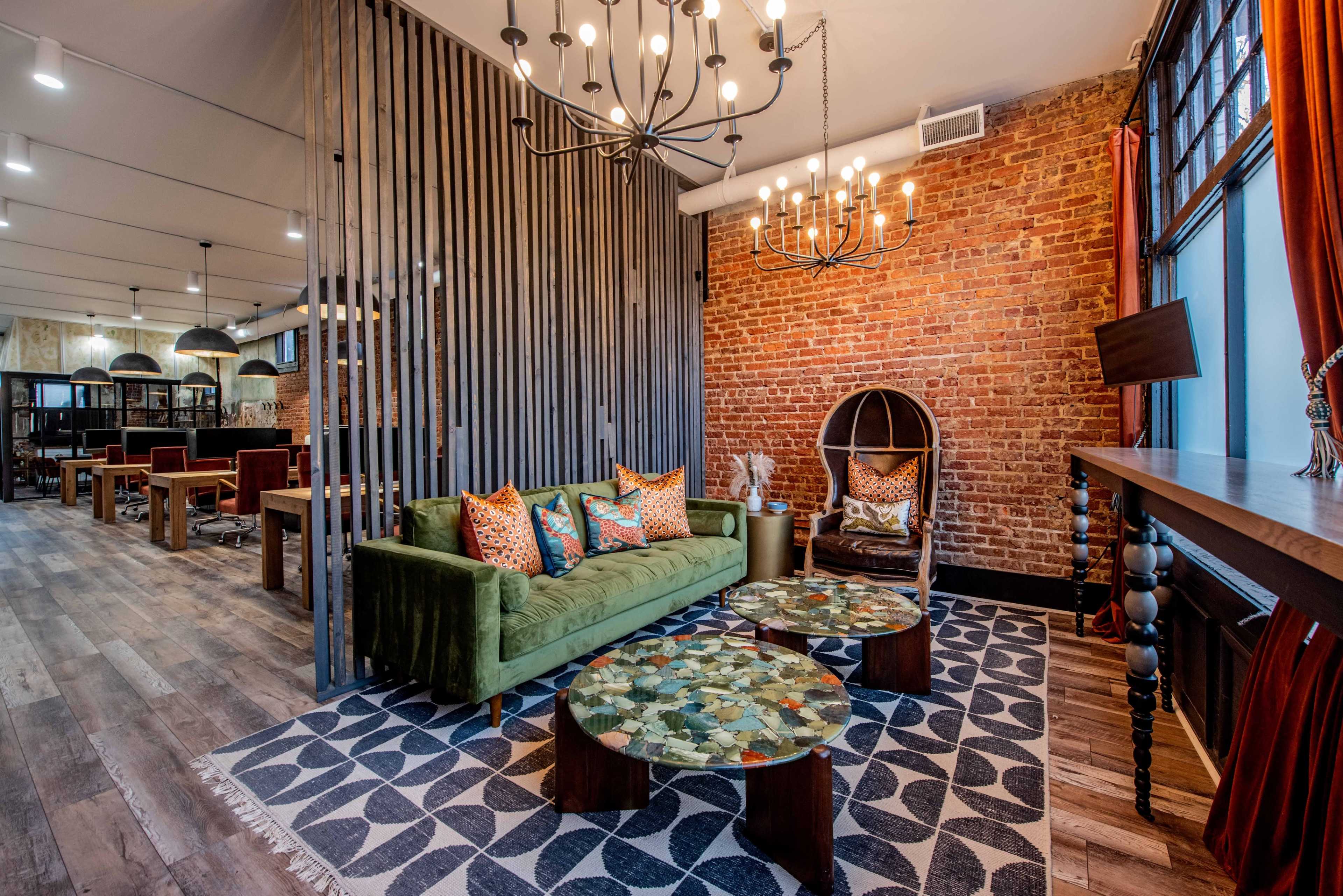 A modern interior featuring a green sofa with patterned pillows, two decorative tables, and a brick wall backdrop, with a chandelier overhead and a workspace area in the background.
