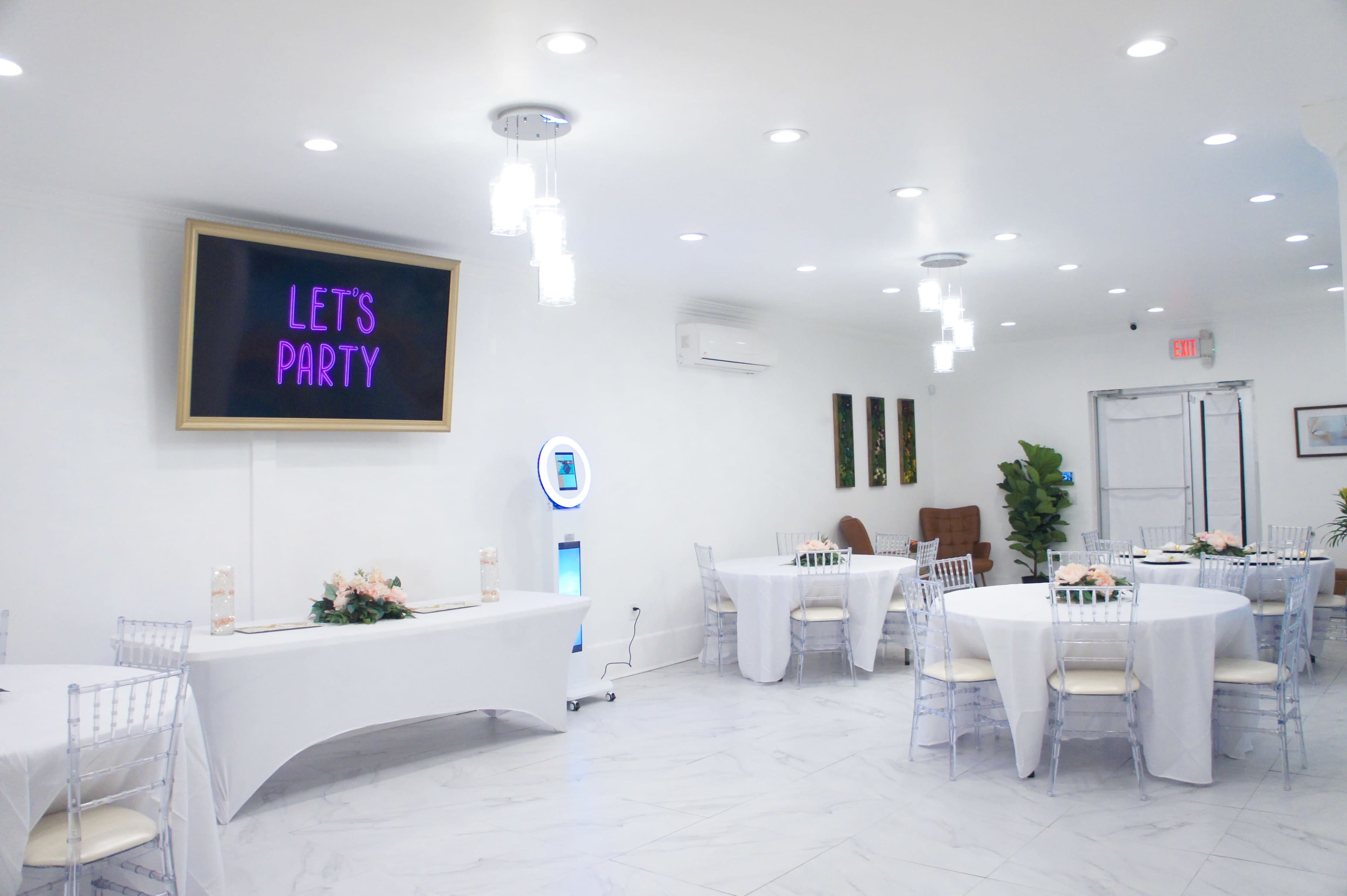 A bright, modern event space with white walls, round tables covered in white linens, and a screen displaying the text "LET'S PARTY."