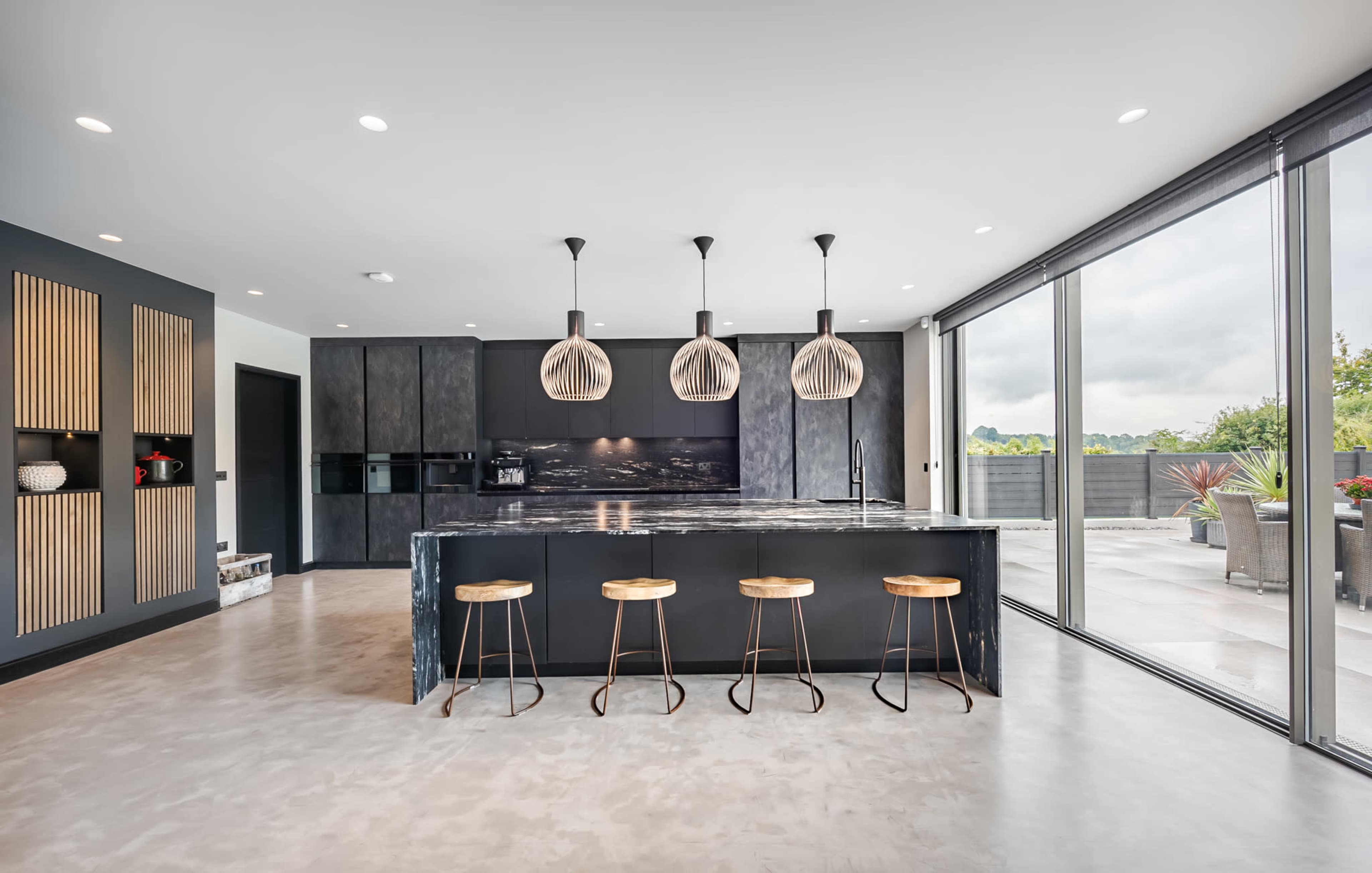 The kitchen features dark cabinetry, a large marble island with three wooden bar stools, and expansive windows overlooking a terrace.