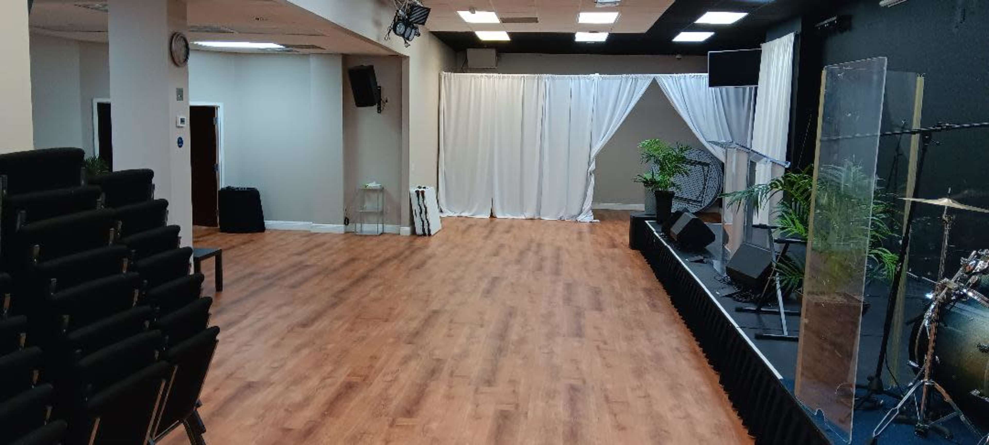 The image shows a spacious room with a wooden floor, a stage with musical instruments, and white curtains in the background.