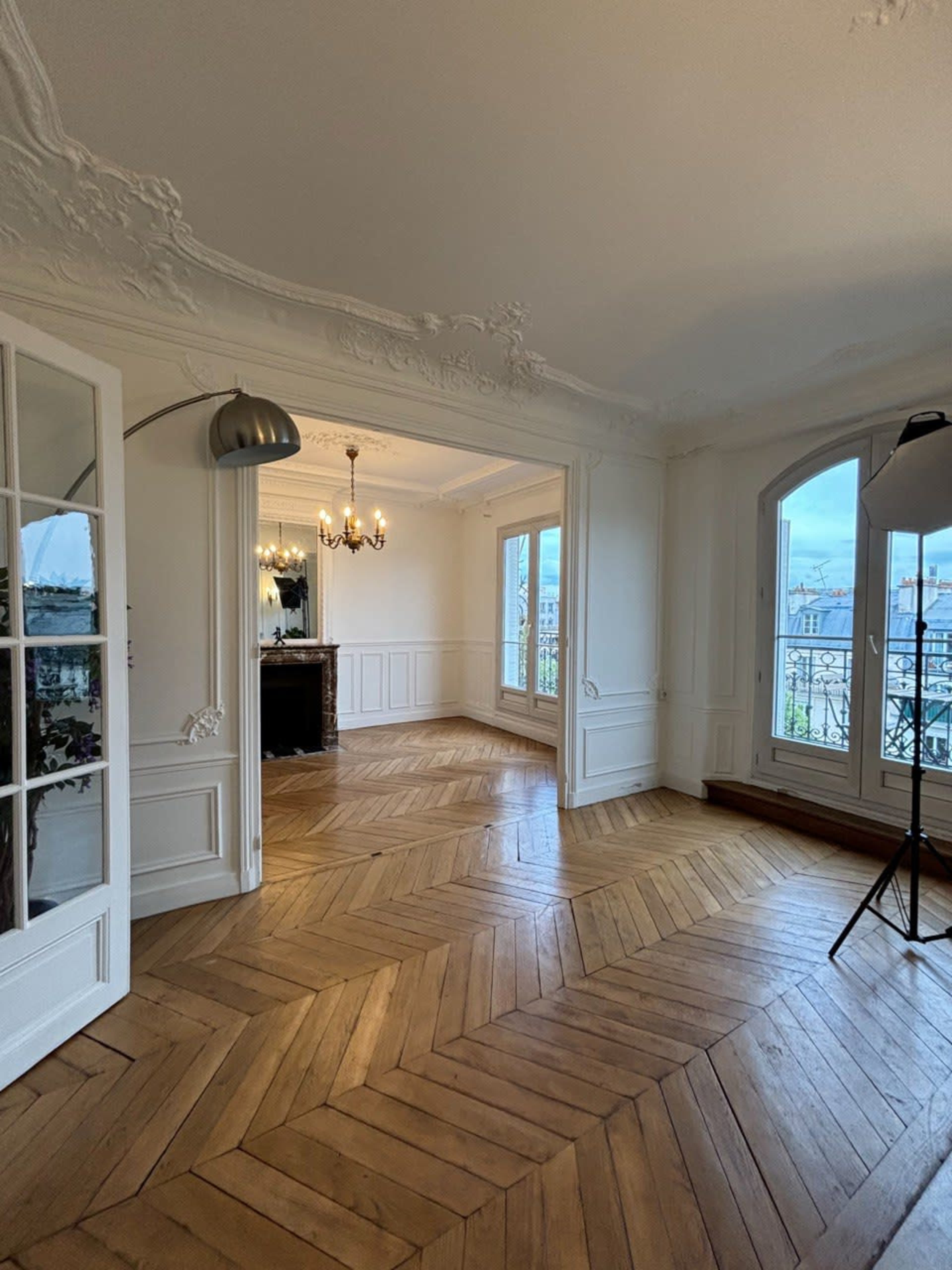 The image shows a spacious room with herringbone-patterned wooden flooring, ornate ceiling details, and large windows providing natural light.
