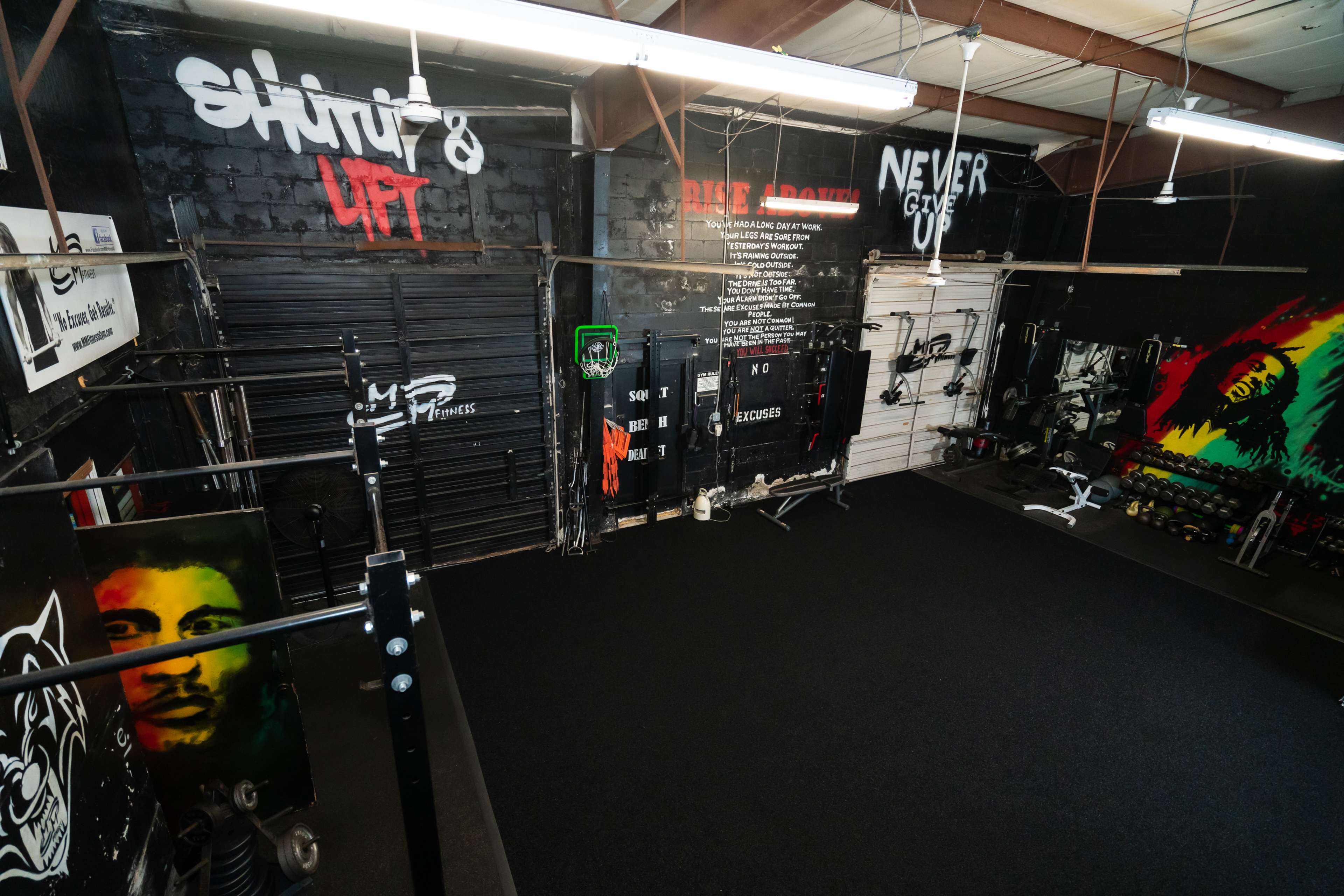 A gym interior with black rubber flooring, walls adorned with various graffiti art, and fitness equipment arranged in the space.