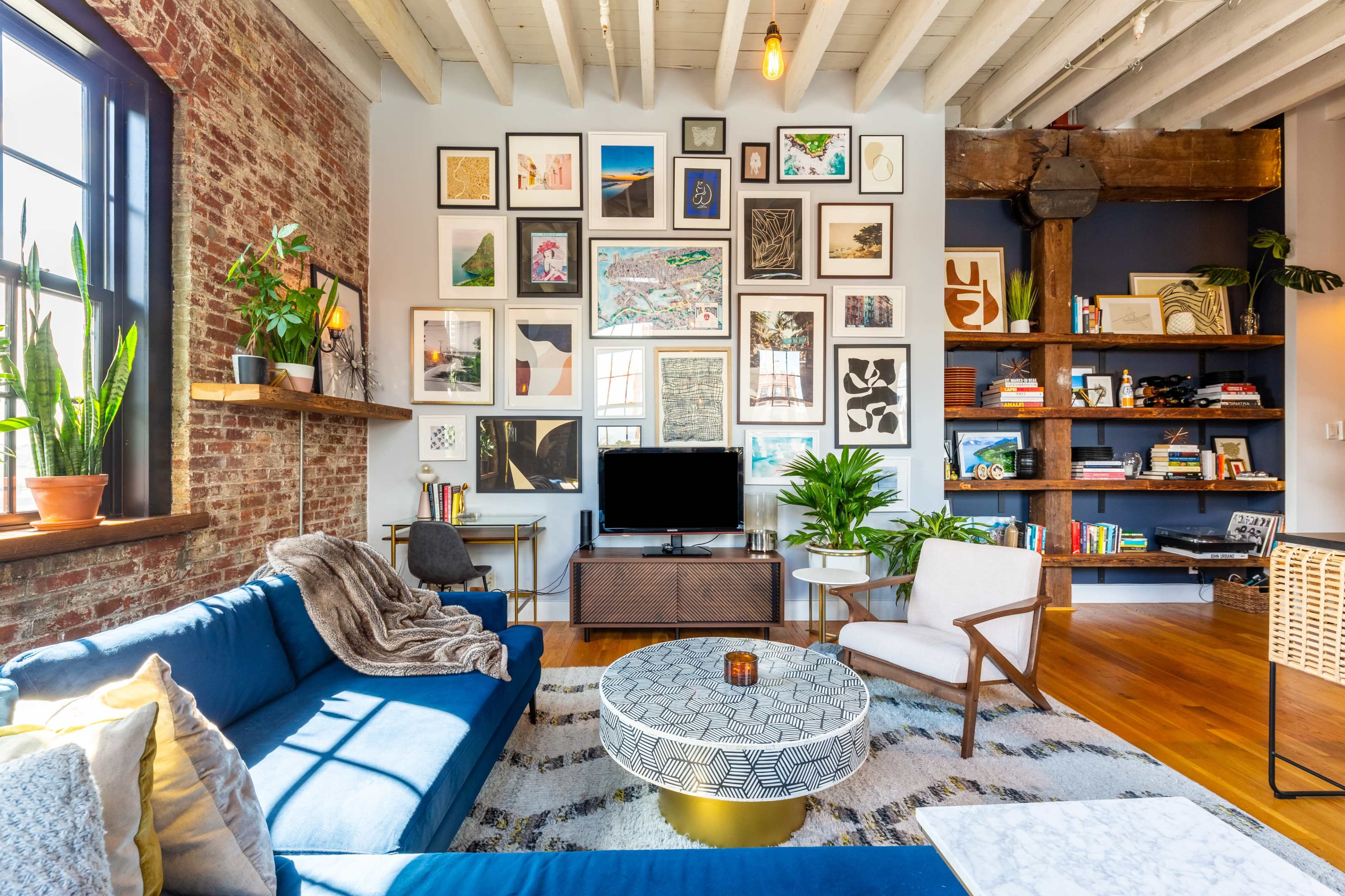 The living room features a blue sofa, a round coffee table, a wall of various framed artworks, and bookshelves filled with books and decorative items.