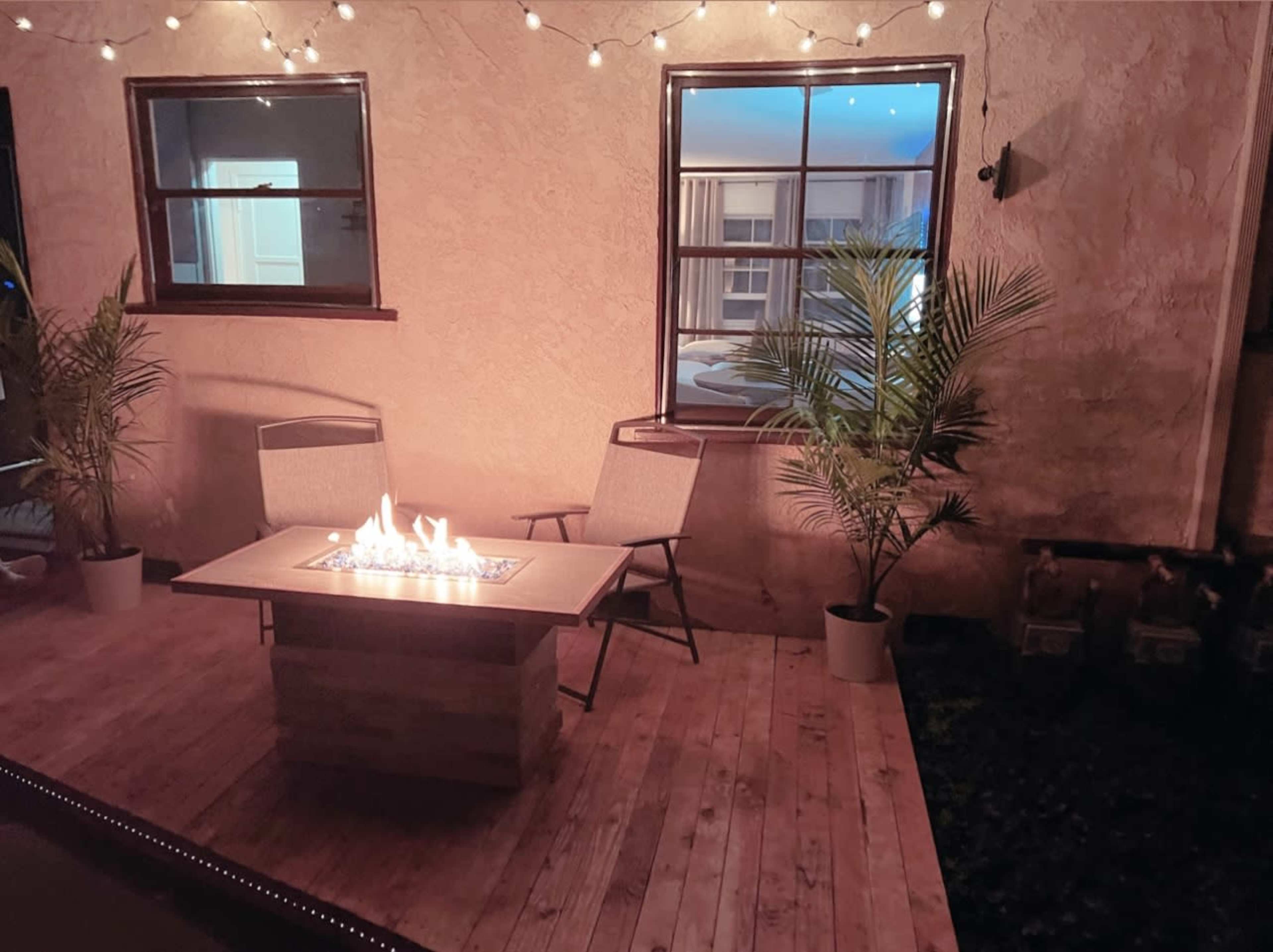 A wooden deck features a fire table surrounded by two chairs and potted plants, with string lights illuminating the area.