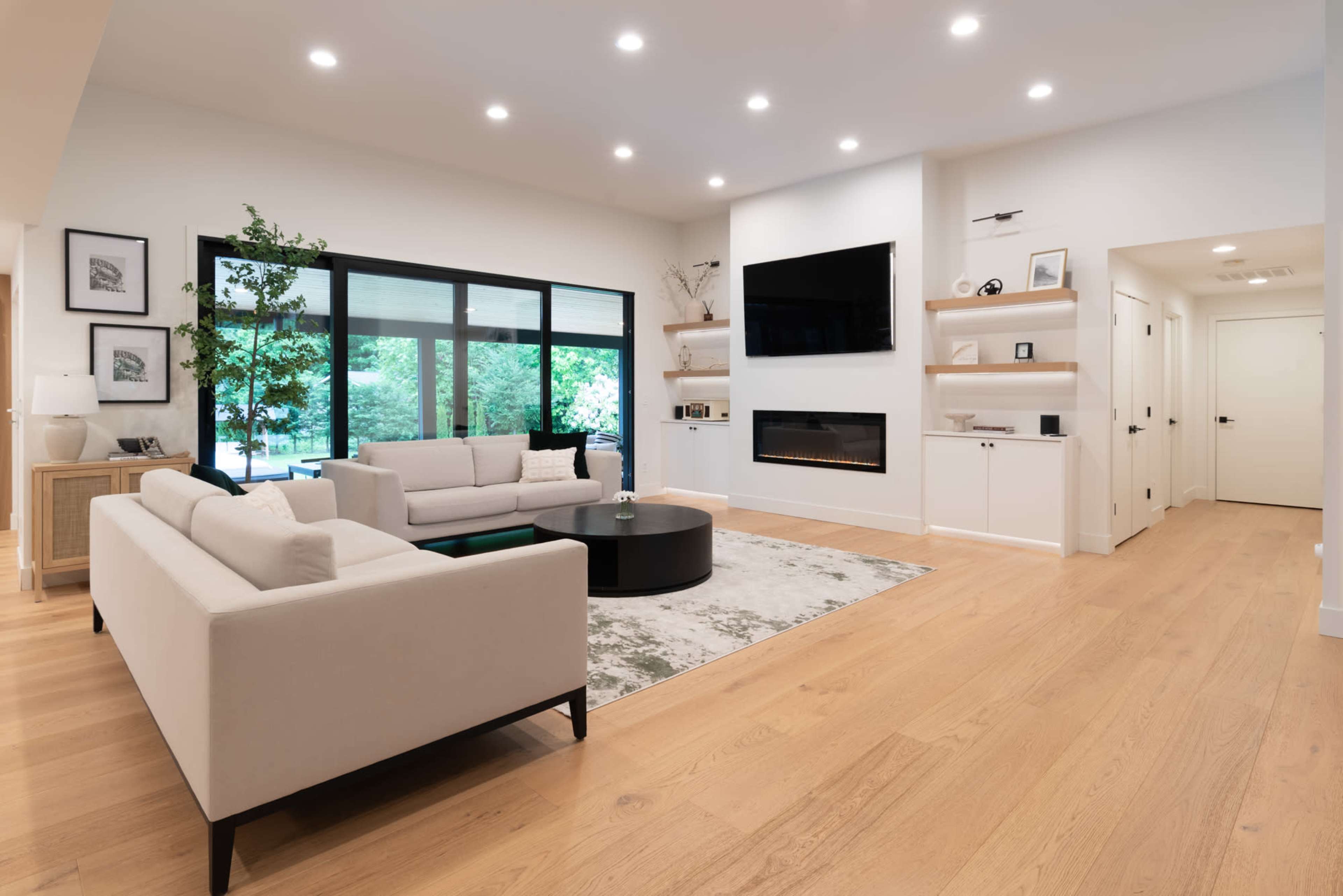 The image shows a modern living room with two light-colored sofas, a round black coffee table, an electric fireplace, and large windows overlooking greenery.