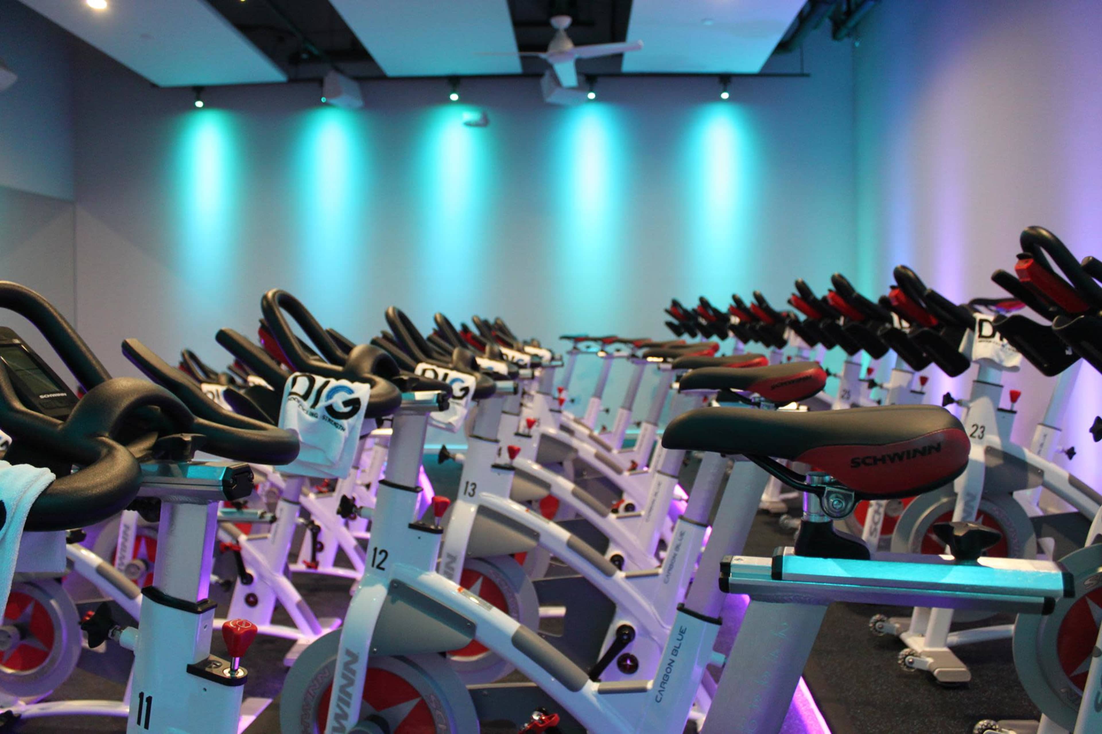 The image shows a row of stationary exercise bikes set up in a brightly lit indoor cycling studio.