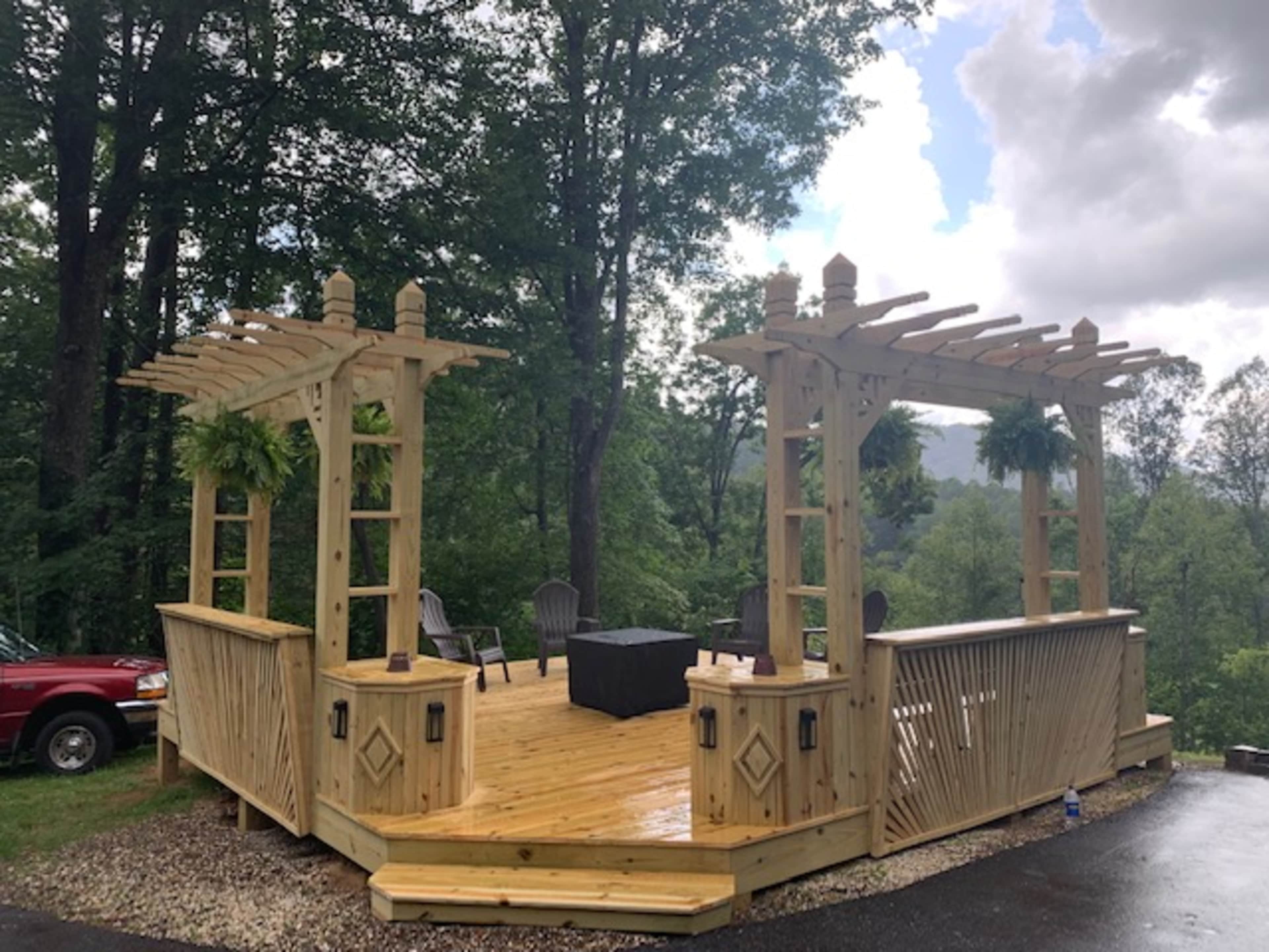 A wooden patio with a pergola design is situated among trees, featuring chairs and a table in a scenic outdoor setting.
