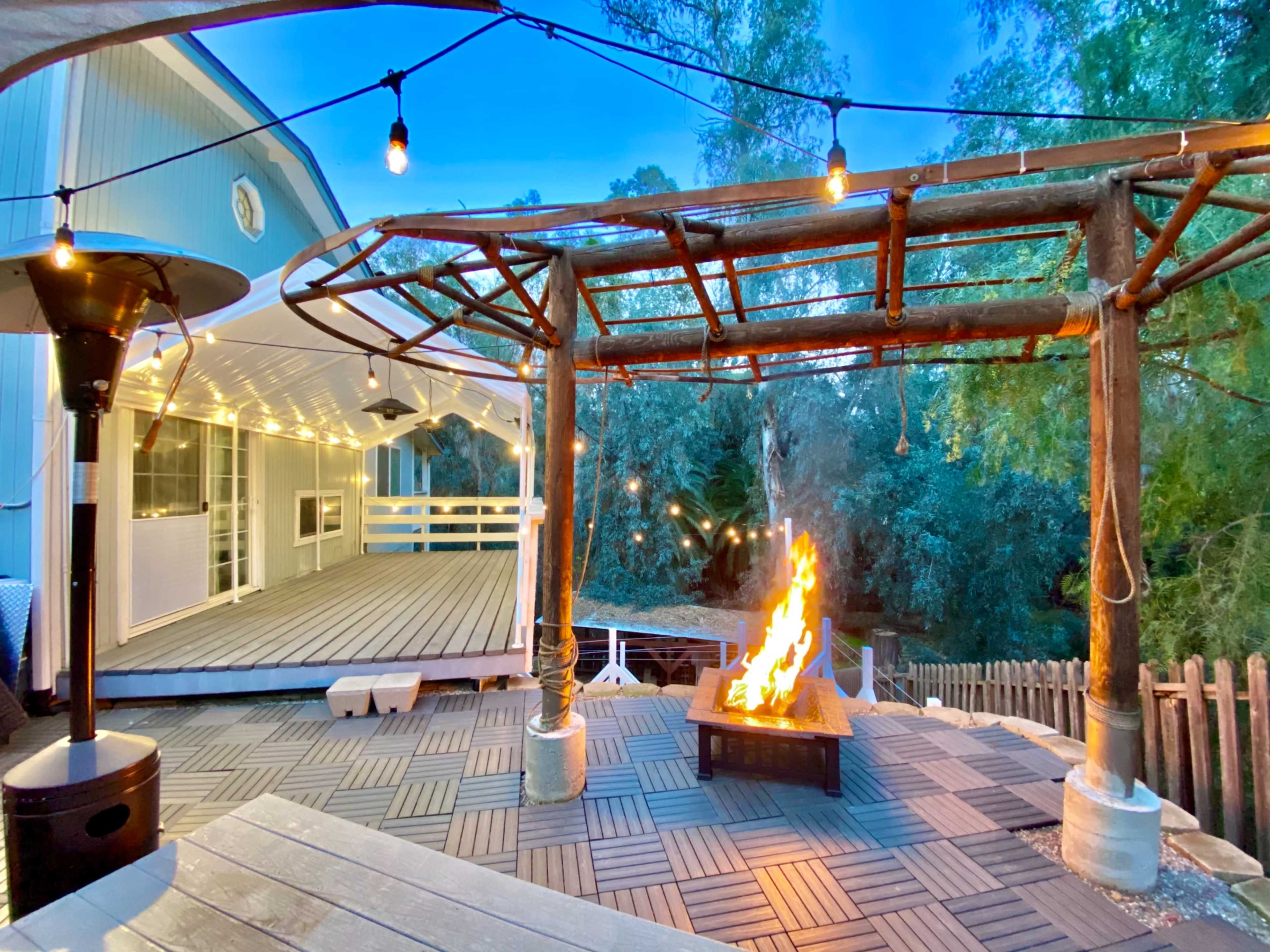 A wooden deck features a fire pit surrounded by seating, with string lights illuminating a pergola in the evening.