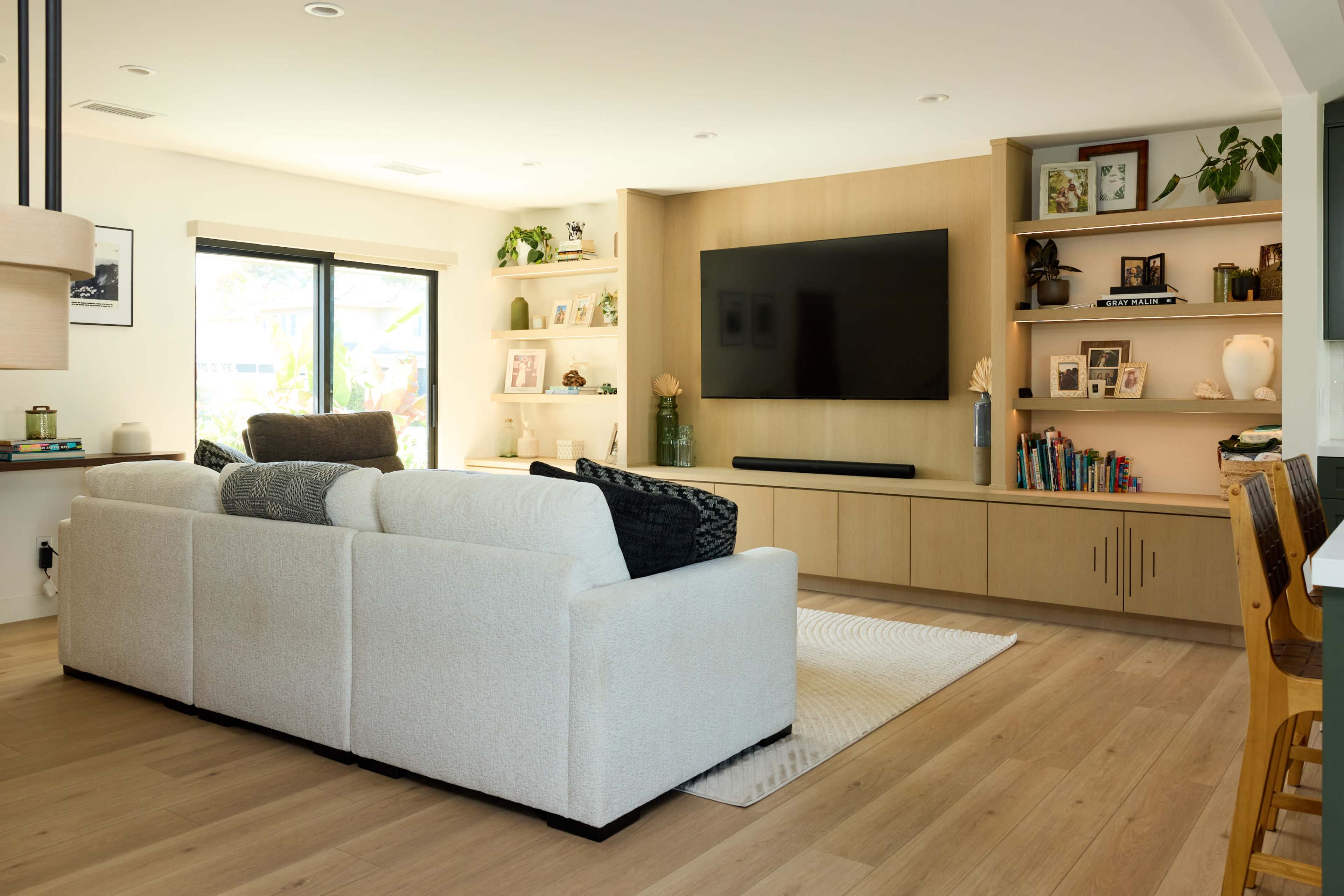 The living room features a light-colored sectional sofa facing a wall-mounted television, flanked by shelves with decorative items and plants.