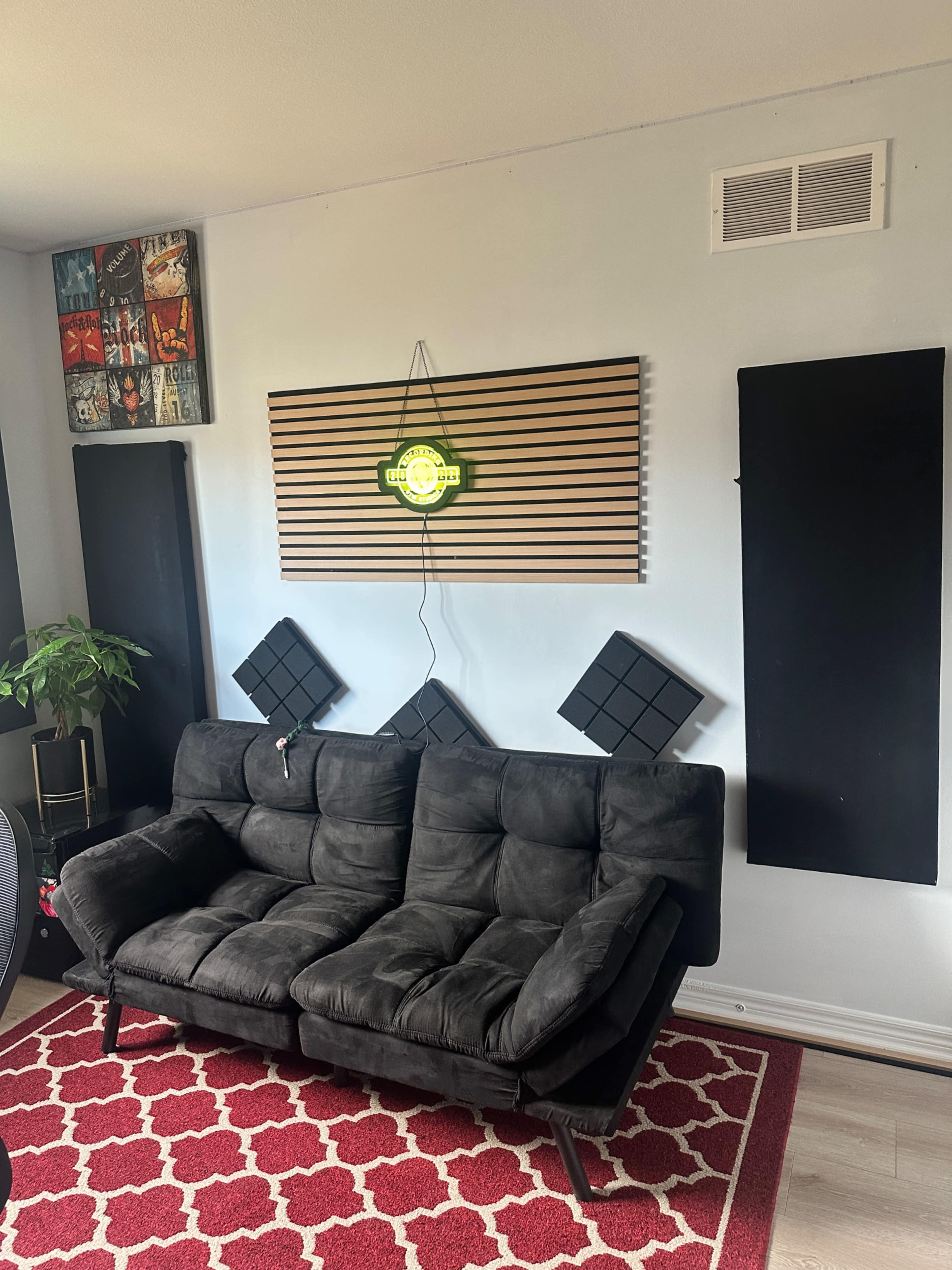 The image shows a black couch against a light blue wall, surrounded by decorative acoustic panels and a green neon sign above a wooden slatted panel.