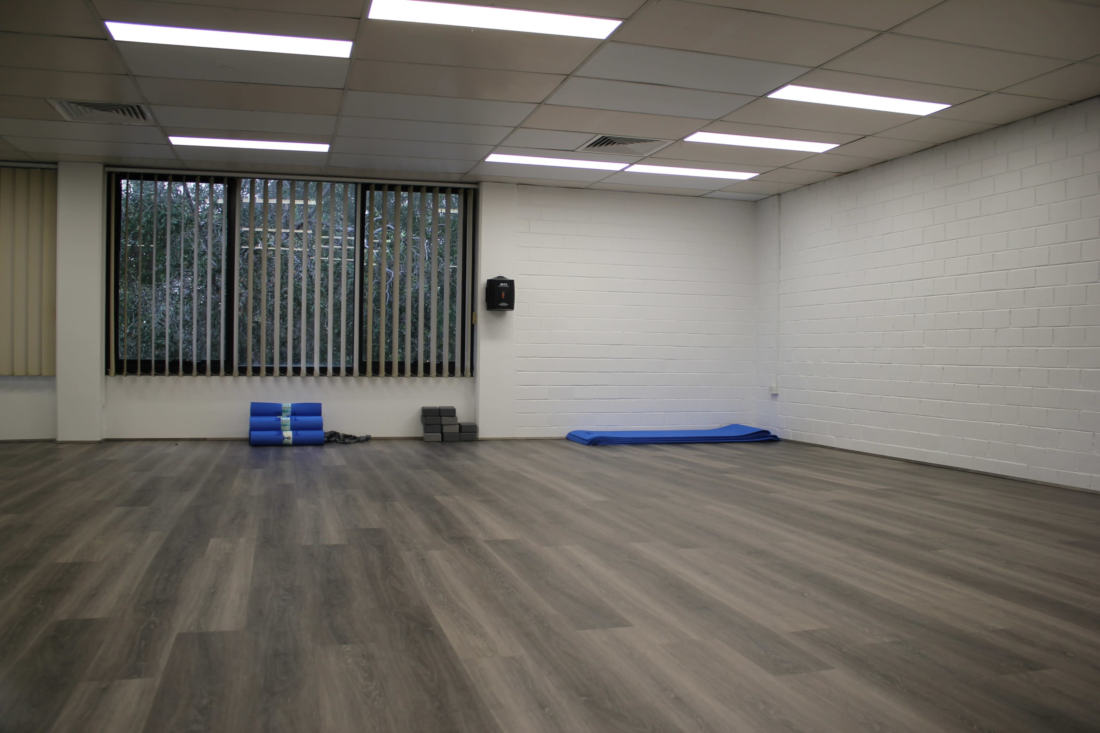 A spacious, empty room with wooden flooring features a blue exercise mat and gym equipment against one wall.