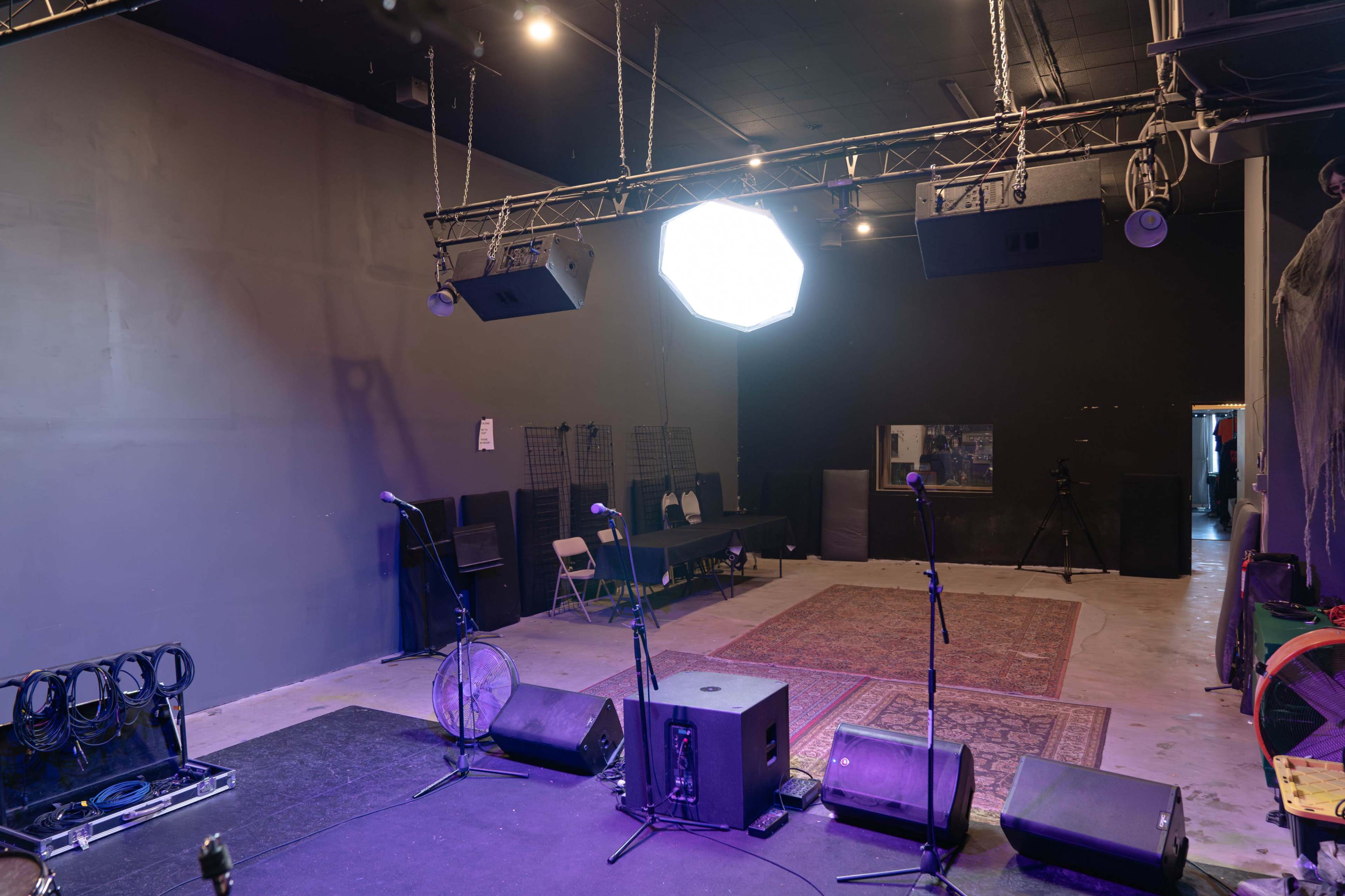 The image shows a performance space with a stage featuring two microphones, speakers, and a rug on the floor, along with chairs and equipment in the background.