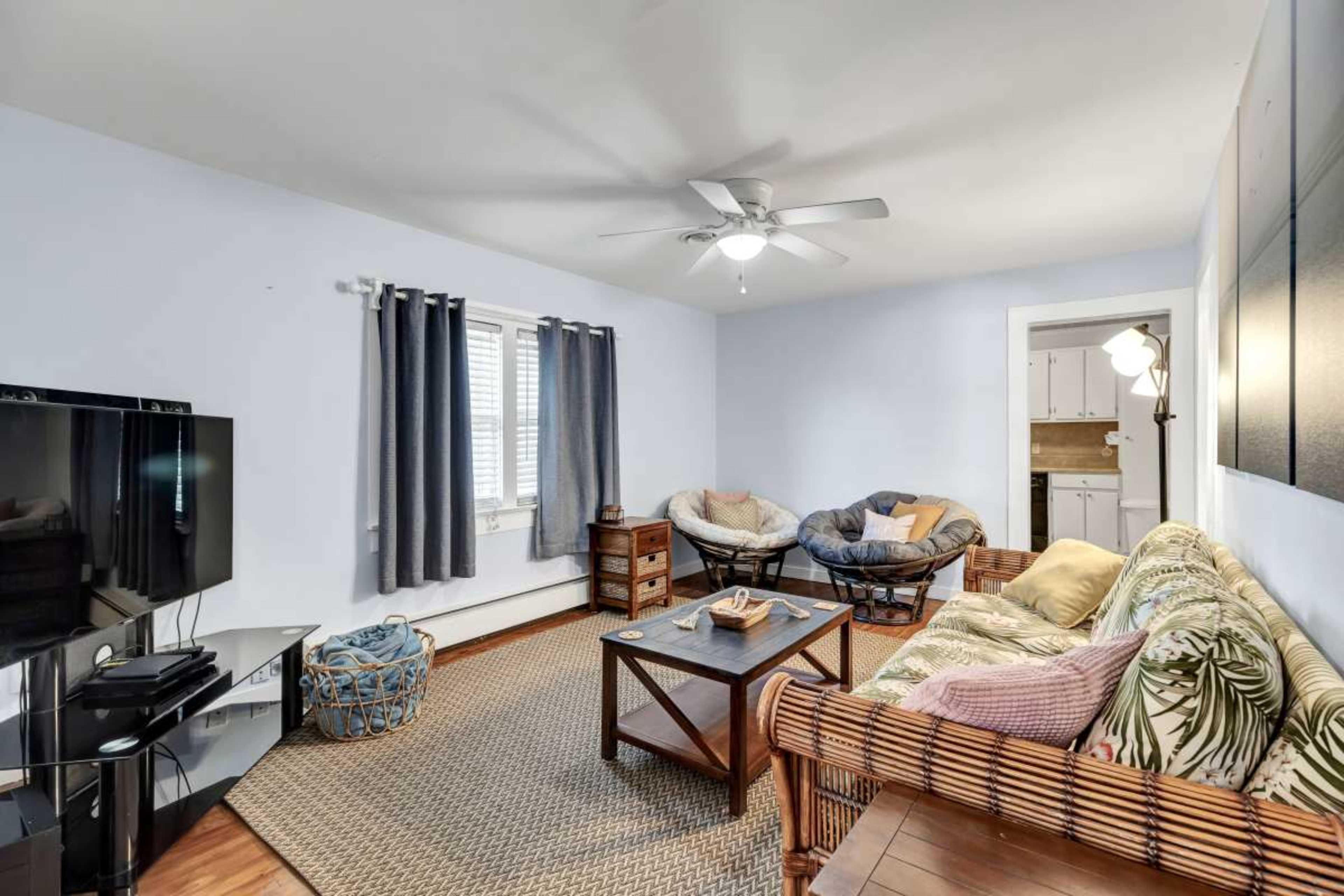The living room features a couch with floral patterns, two cushioned chairs, a coffee table, a television unit, and windows dressed with curtains, creating a cozy atmosphere.