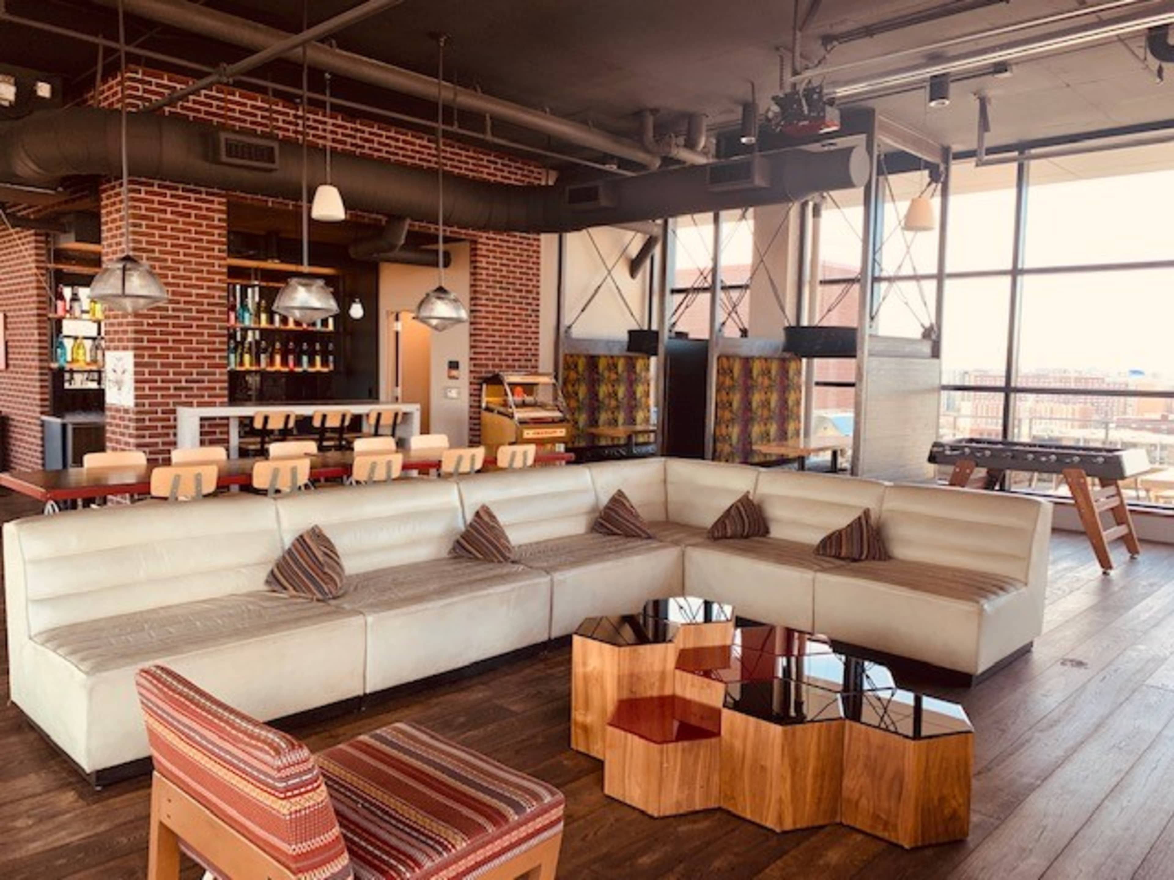 A modern lounge area with a large white sectional sofa, wooden coffee tables, and a bar in the background featuring shelves of bottles.