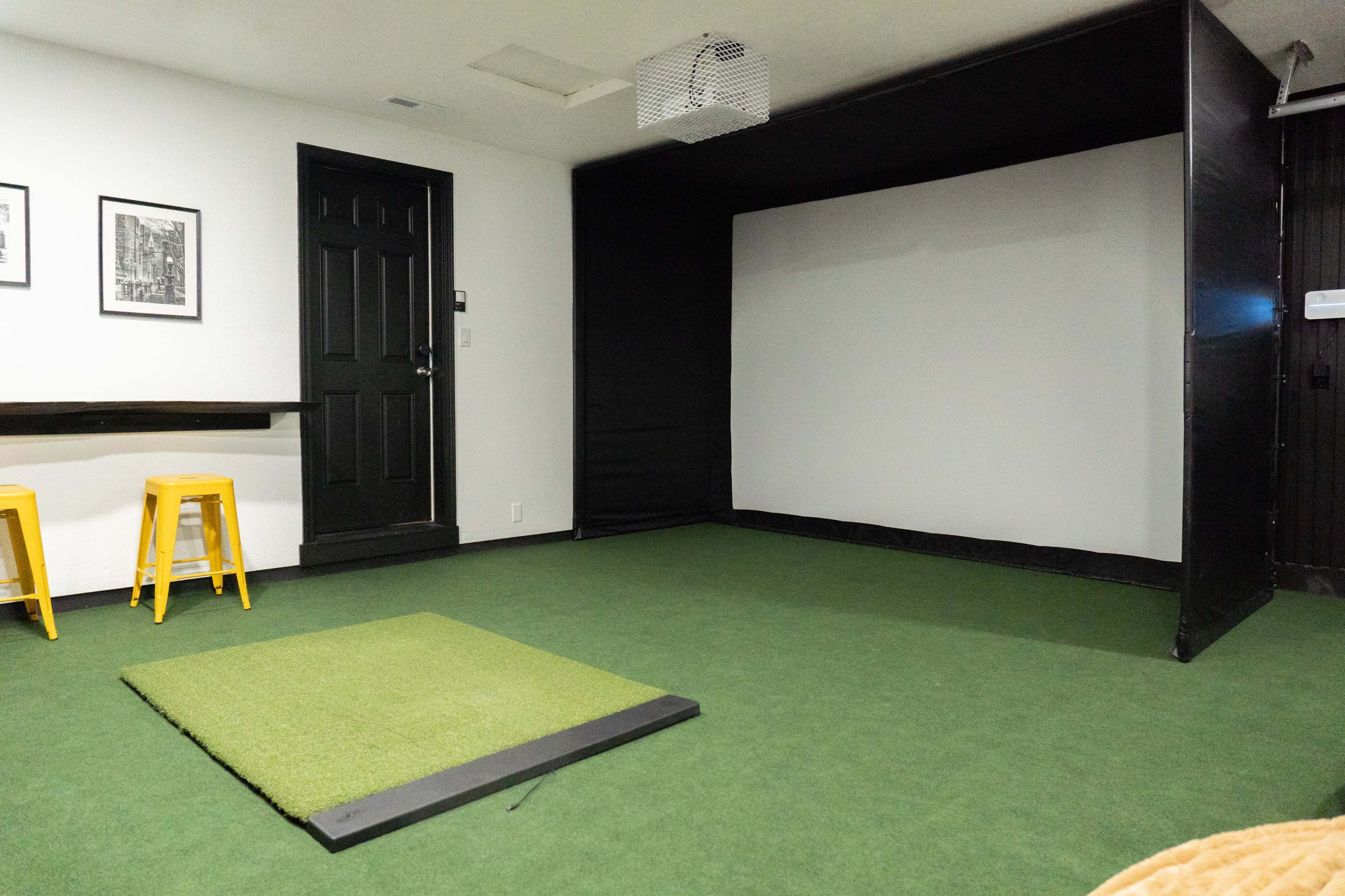 An indoor room with a green carpeted floor, featuring a hitting mat in front of a large black projection screen and a door on one side.
