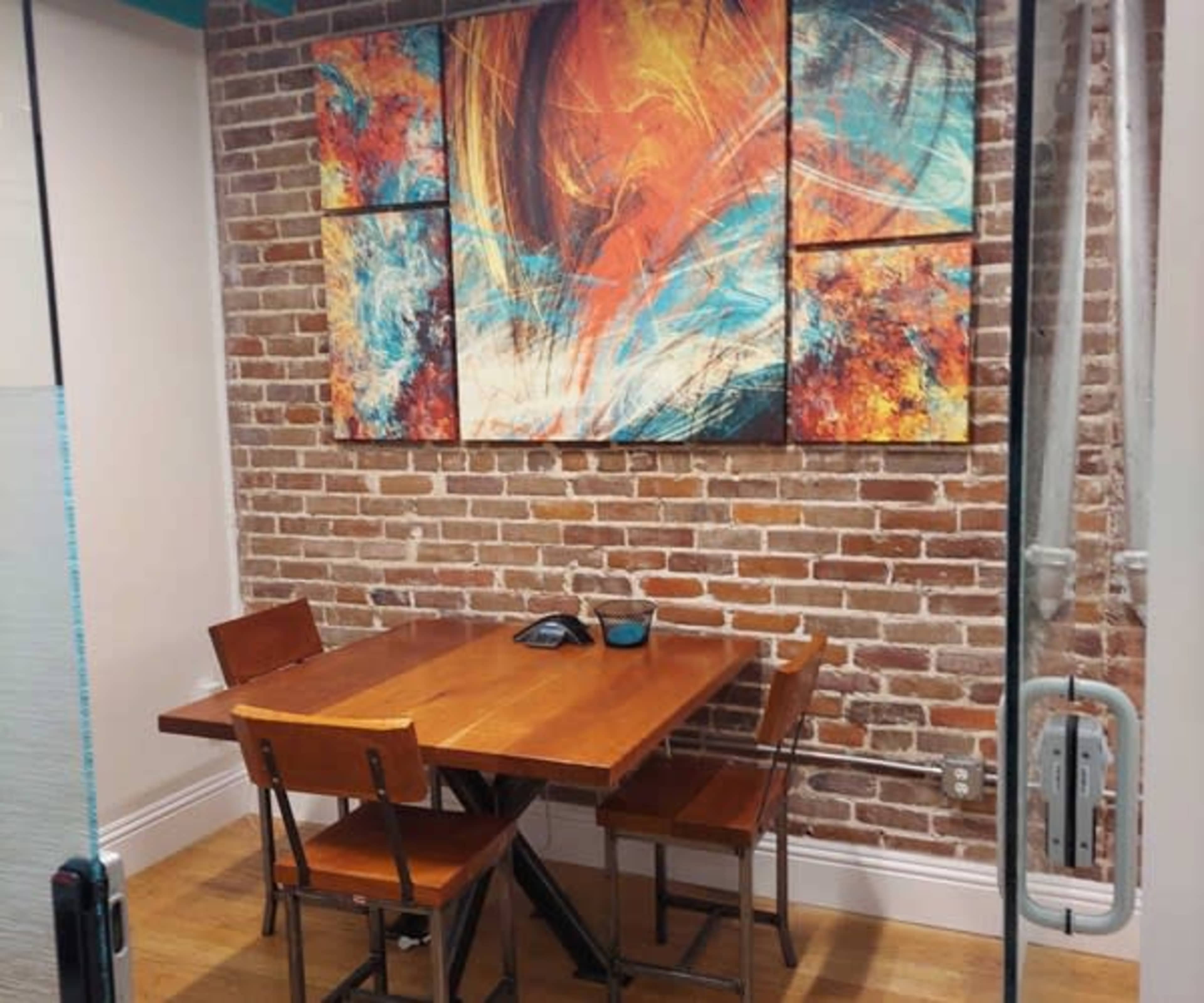 The image shows a small meeting room with a wooden table and four chairs set against a brick wall, featuring an abstract painting above the table.