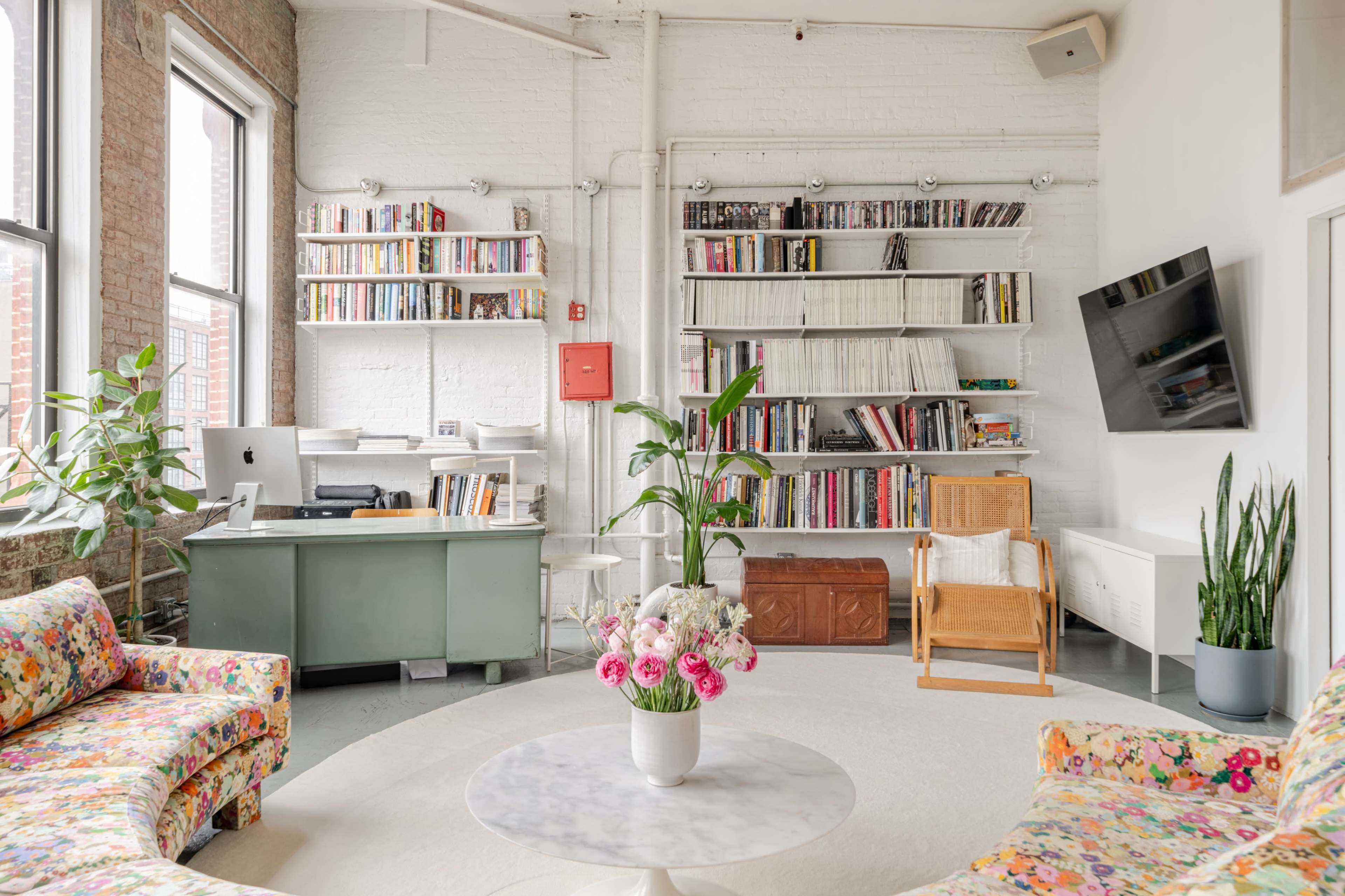 The image shows a bright, modern room with two floral-patterned sofas, a round coffee table, a desk with a computer, shelves filled with books, and tall indoor plants.
