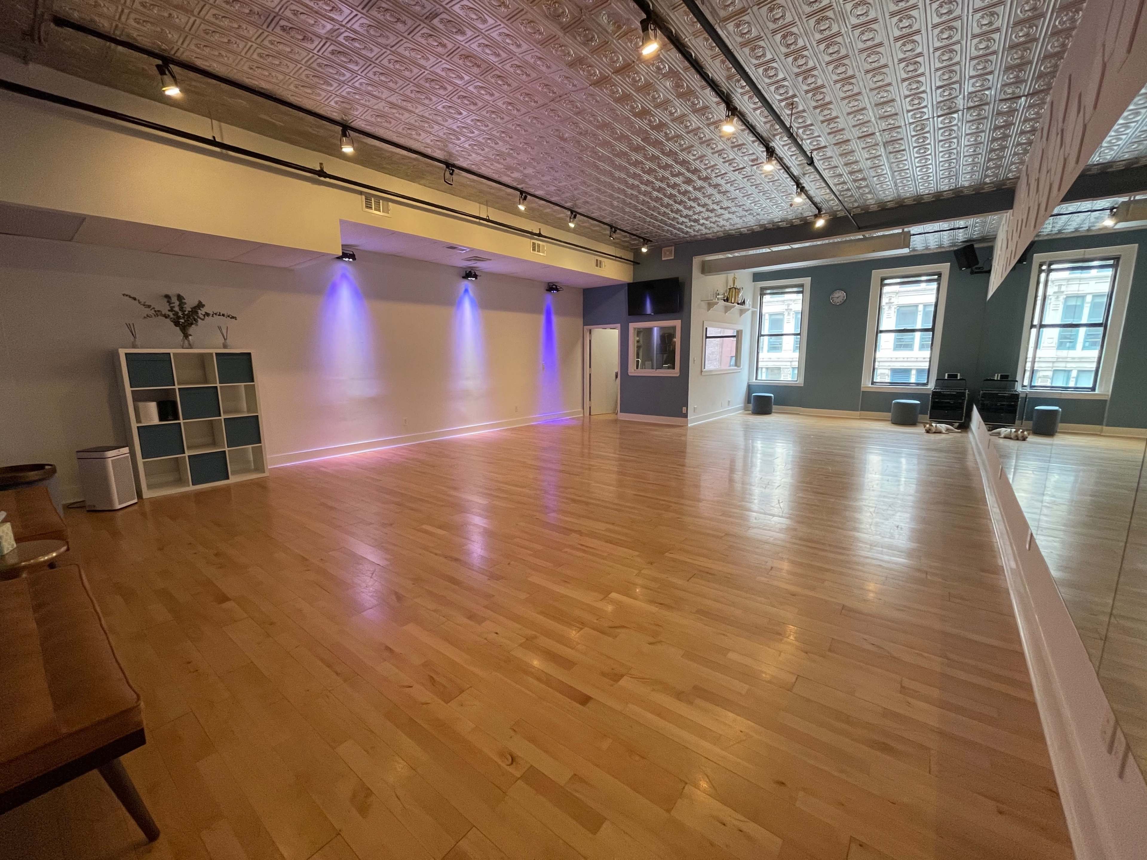 The image shows a spacious studio with wooden flooring, wall-mounted mirrors, and soft lighting.