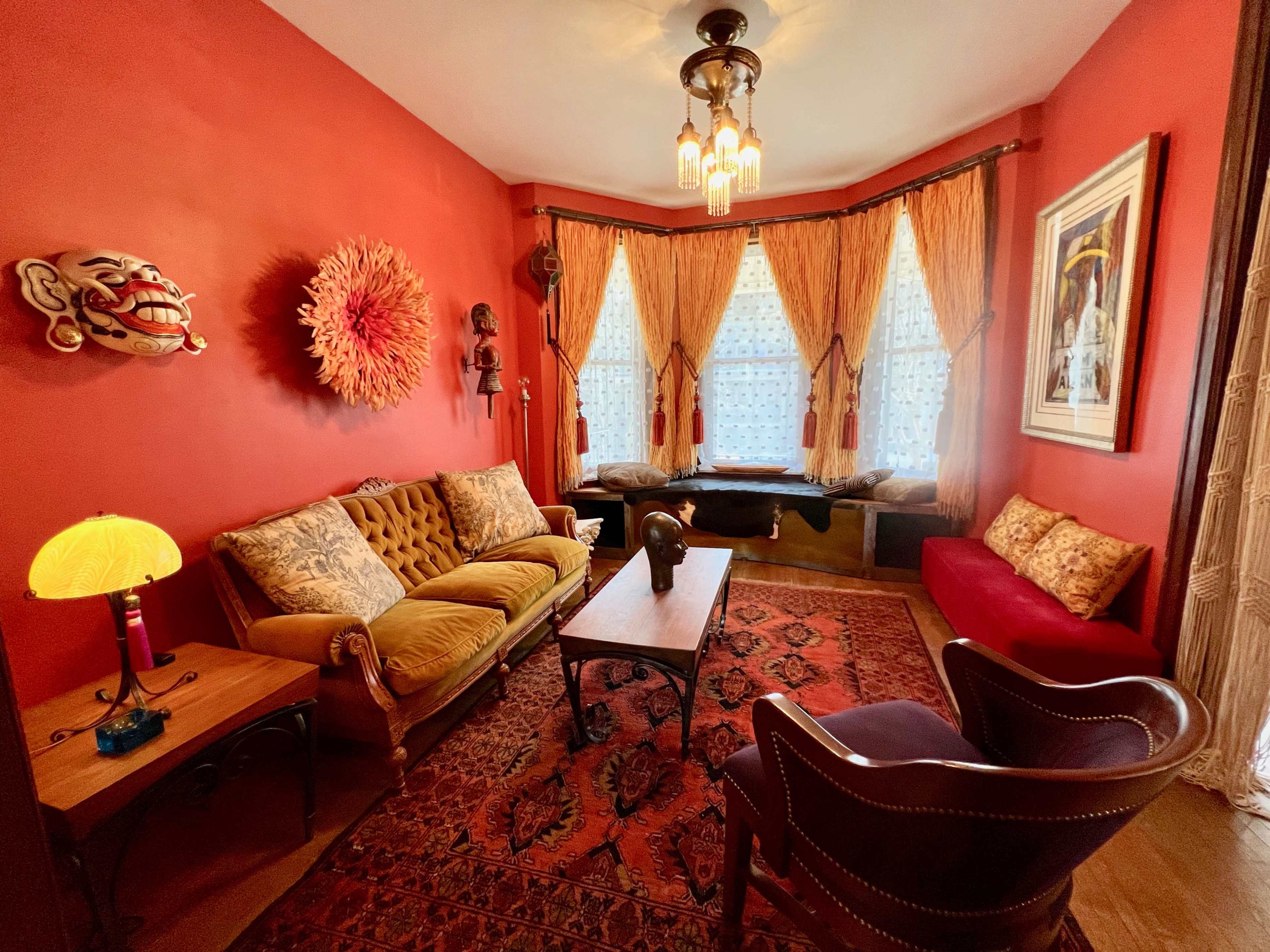 The image shows a cozy living room with red walls, a plush sofa, a wooden coffee table, and large windows draped with beige curtains.