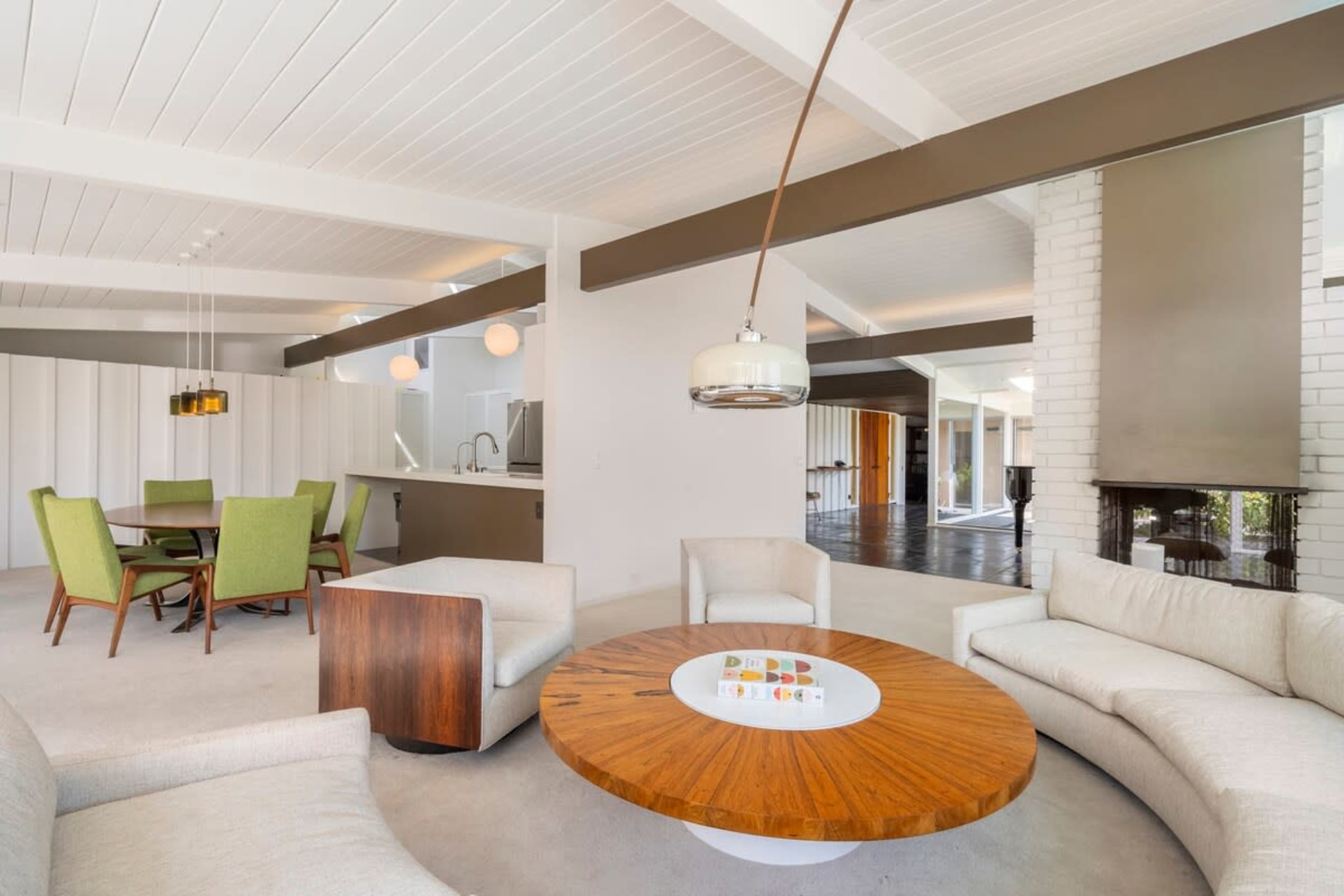 The image shows a modern living space featuring a circular wooden coffee table, light-colored sectional sofas, a dining area with green chairs, and a kitchen visible in the background.