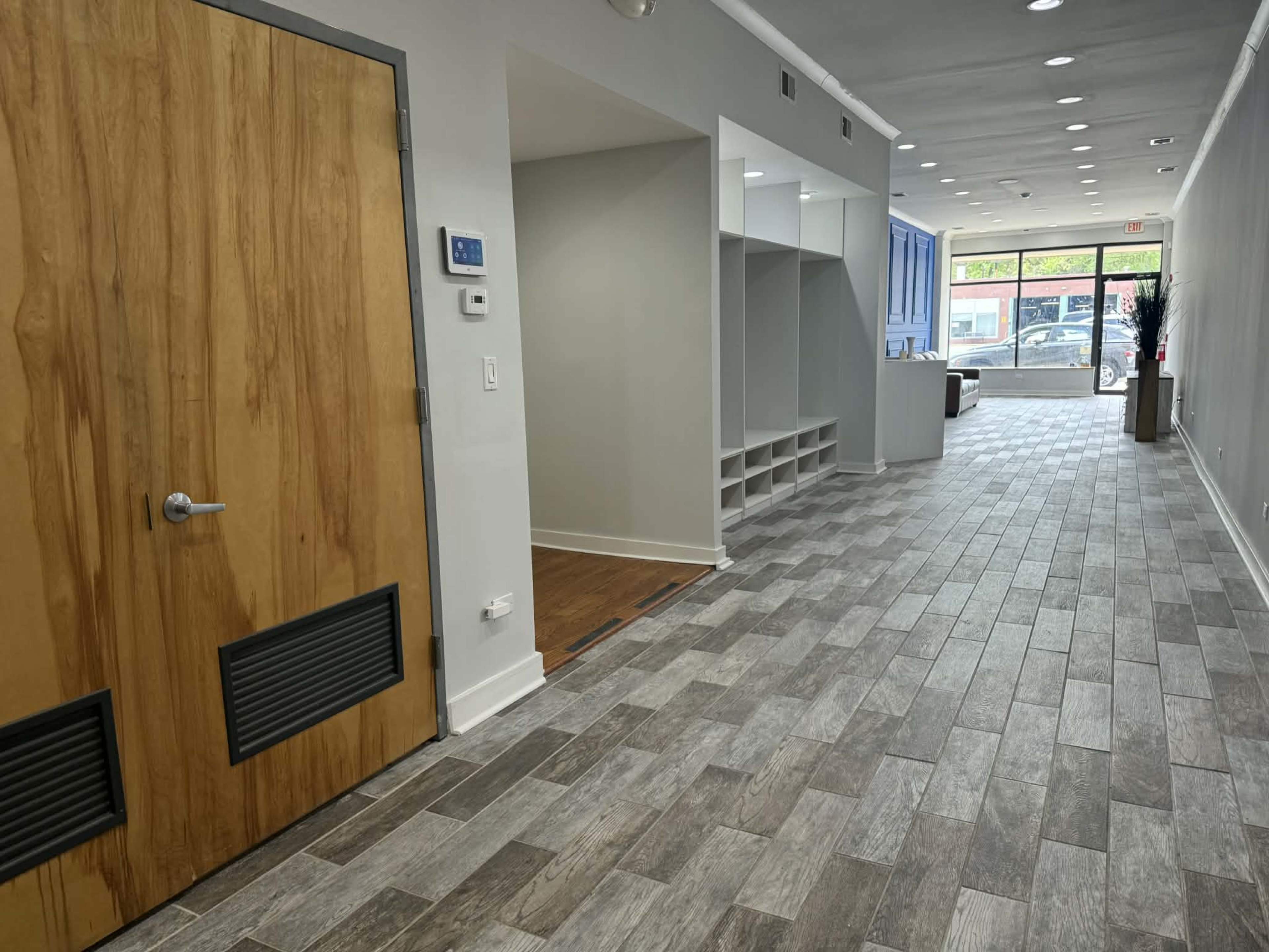 The image shows a modern hallway with wooden flooring, a large door on the left, and a row of built-in shelves along the right side, leading to a storefront at the far end.