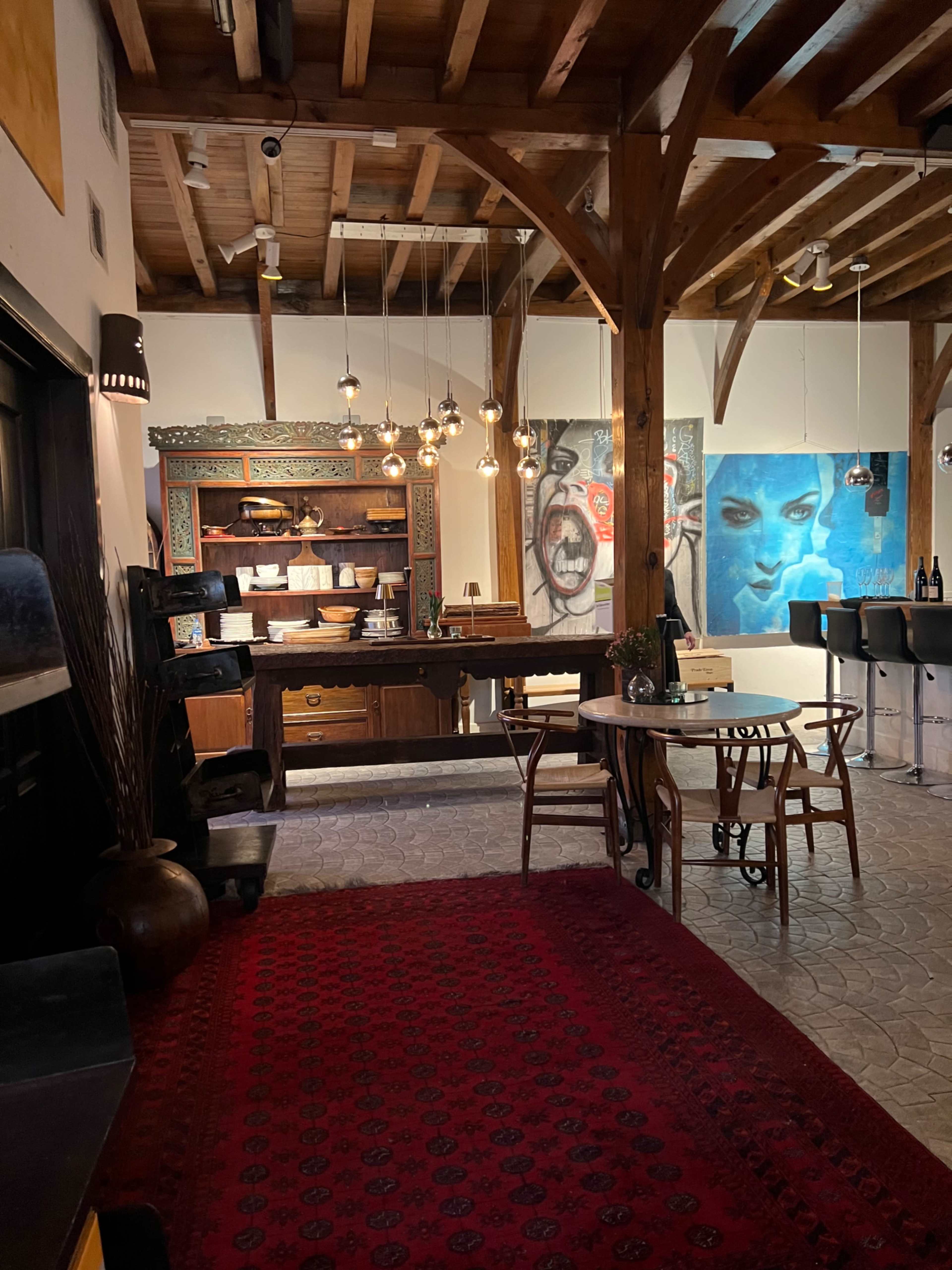The image shows a spacious interior with wooden beams, a rustic table, several chairs, and artworks displayed on the walls.