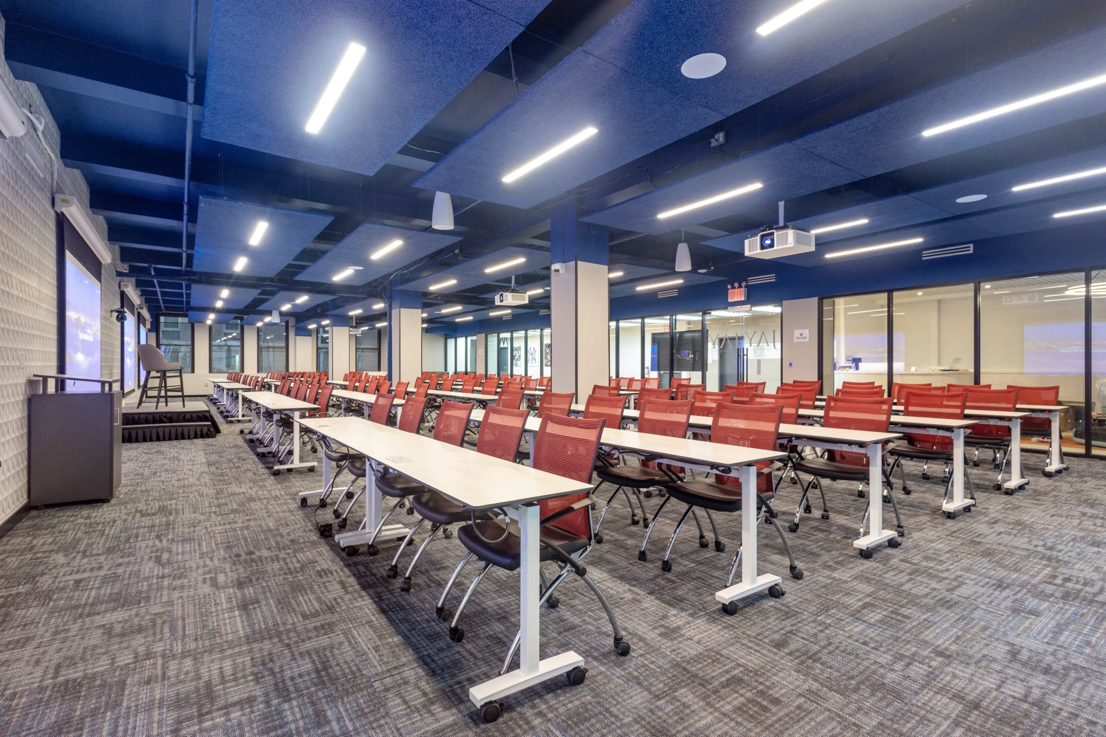 The image shows a modern classroom setup with rows of red chairs and white tables arranged for a lecture or presentation.