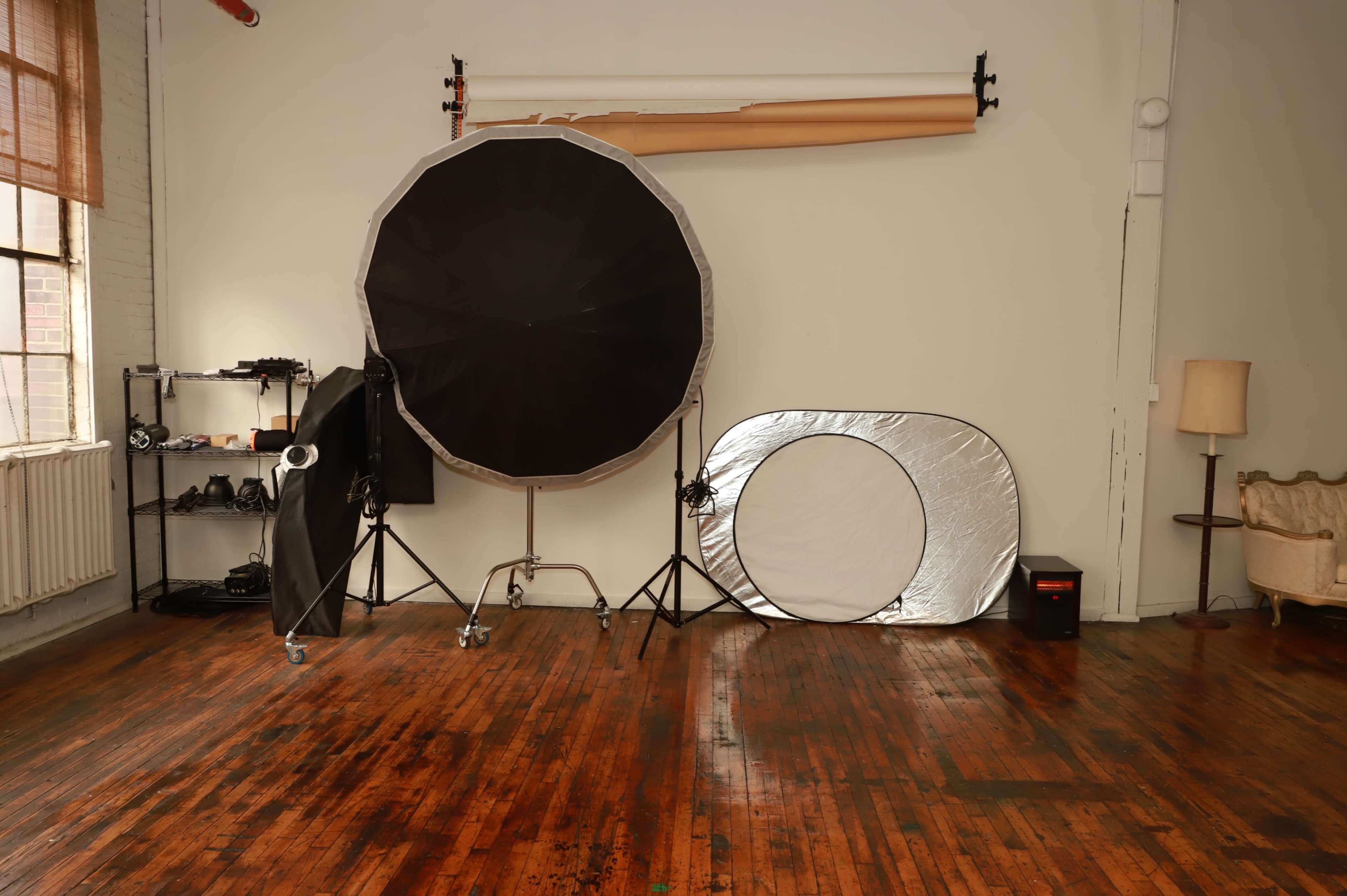 A photography studio features a large softbox, a reflector, and various equipment arranged against hardwood floors and white walls.