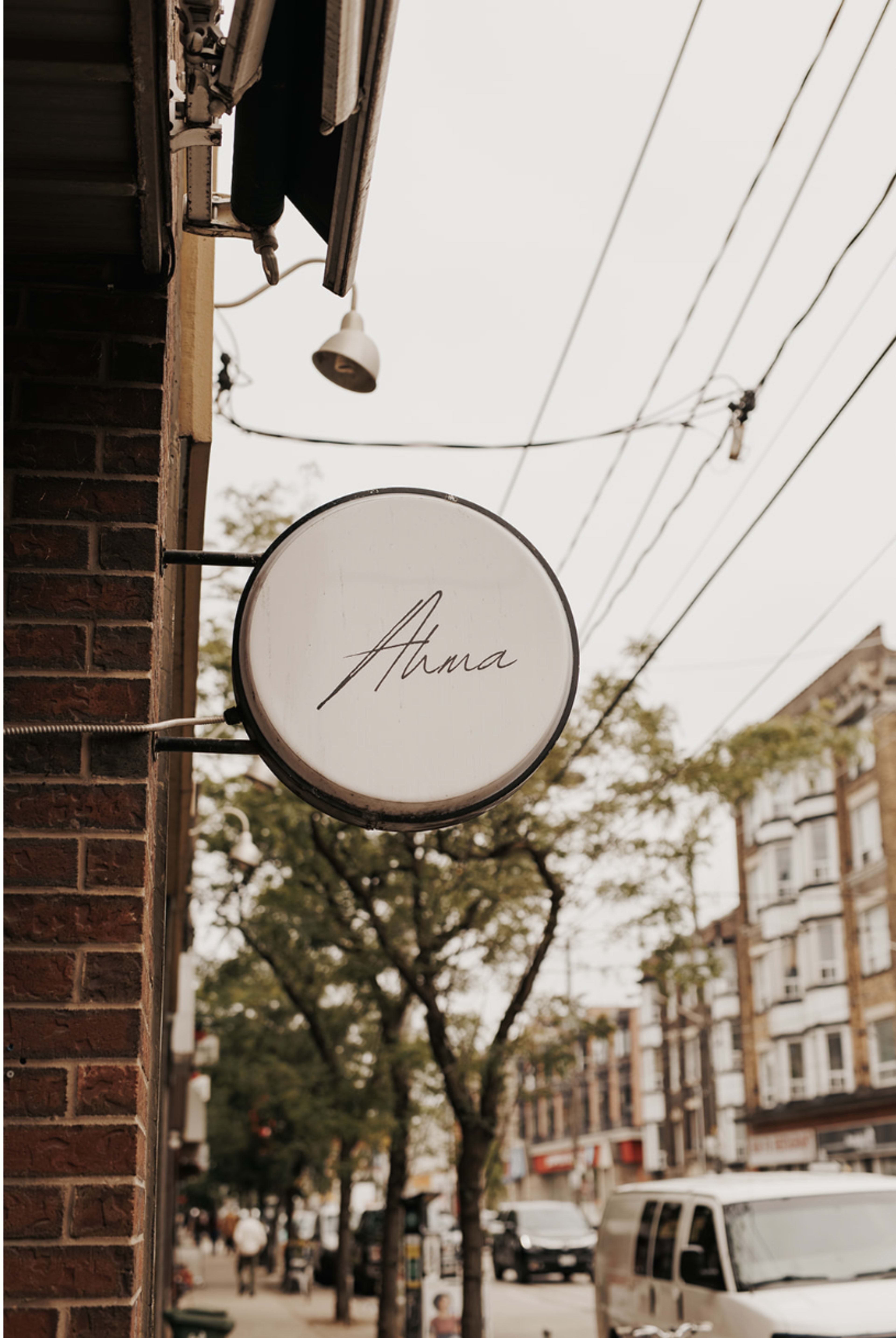 A round sign with the name "Anna" is suspended from a brick building, with power lines and trees visible in the background.