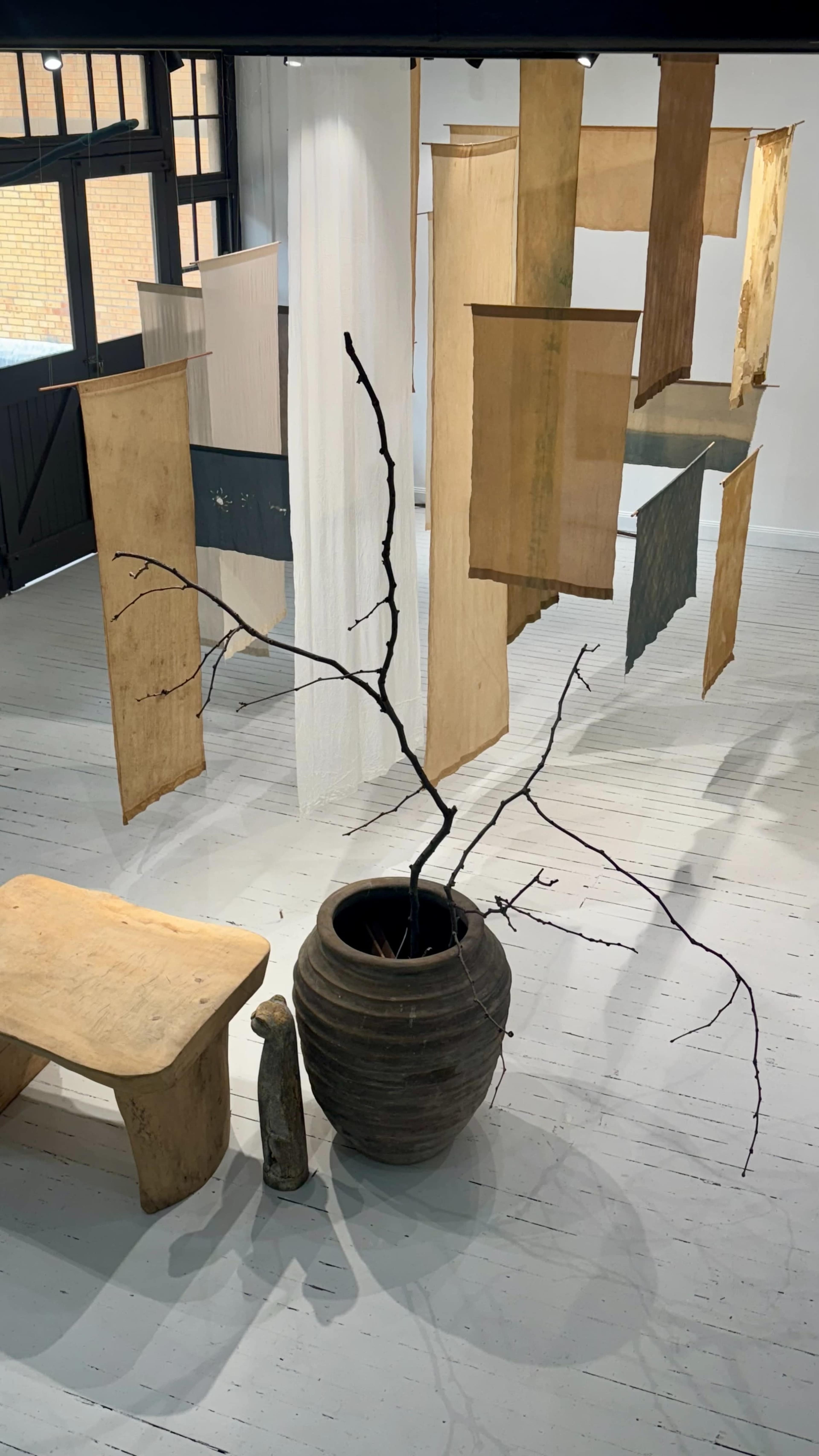 The scene features a minimalist gallery space with hanging fabric panels and an art installation consisting of a large clay pot, a wooden bench, and a bare branch.