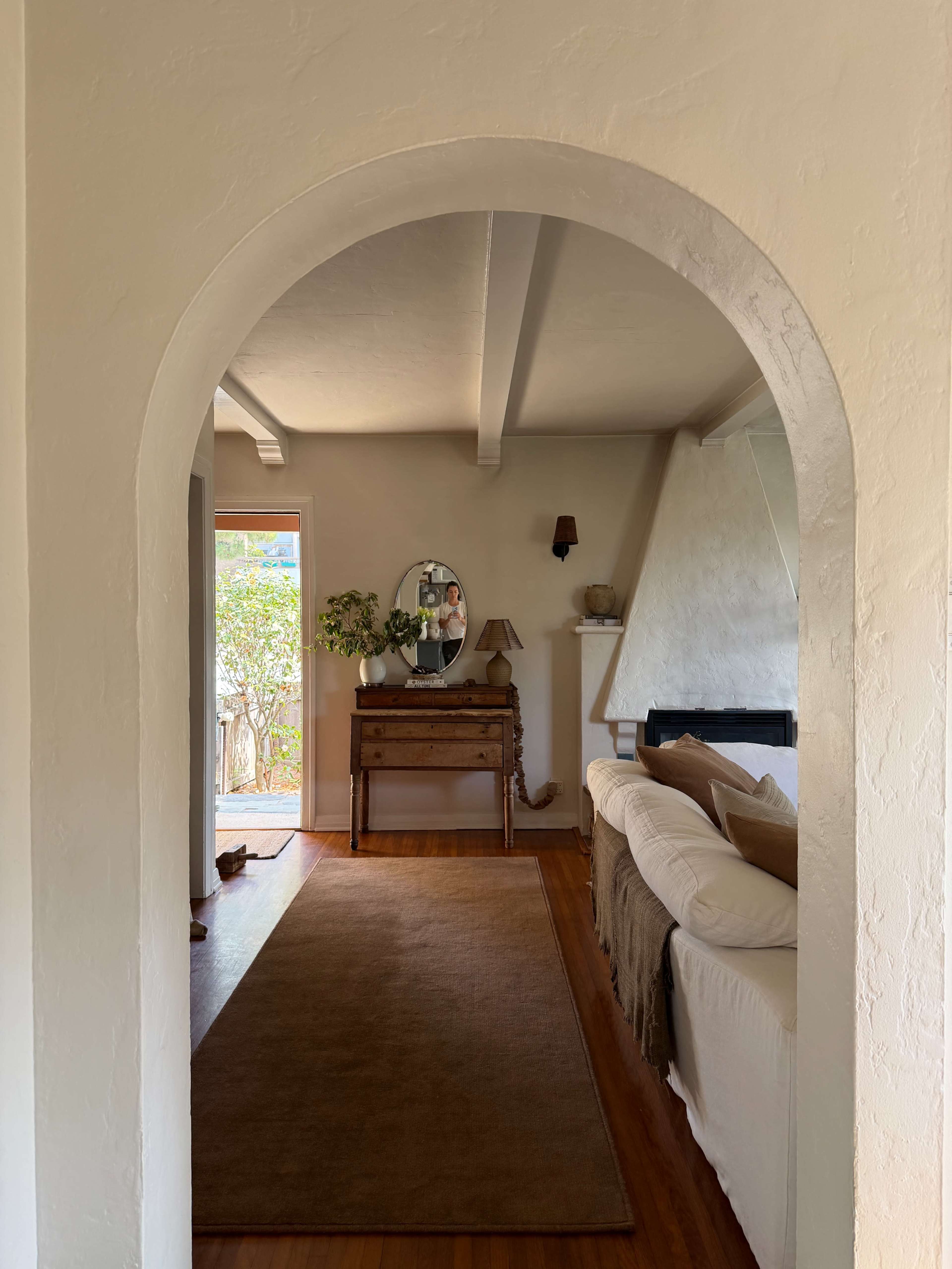 A view through an arched doorway into a living space featuring a sofa, wooden furniture, and natural light spilling in from a nearby door.