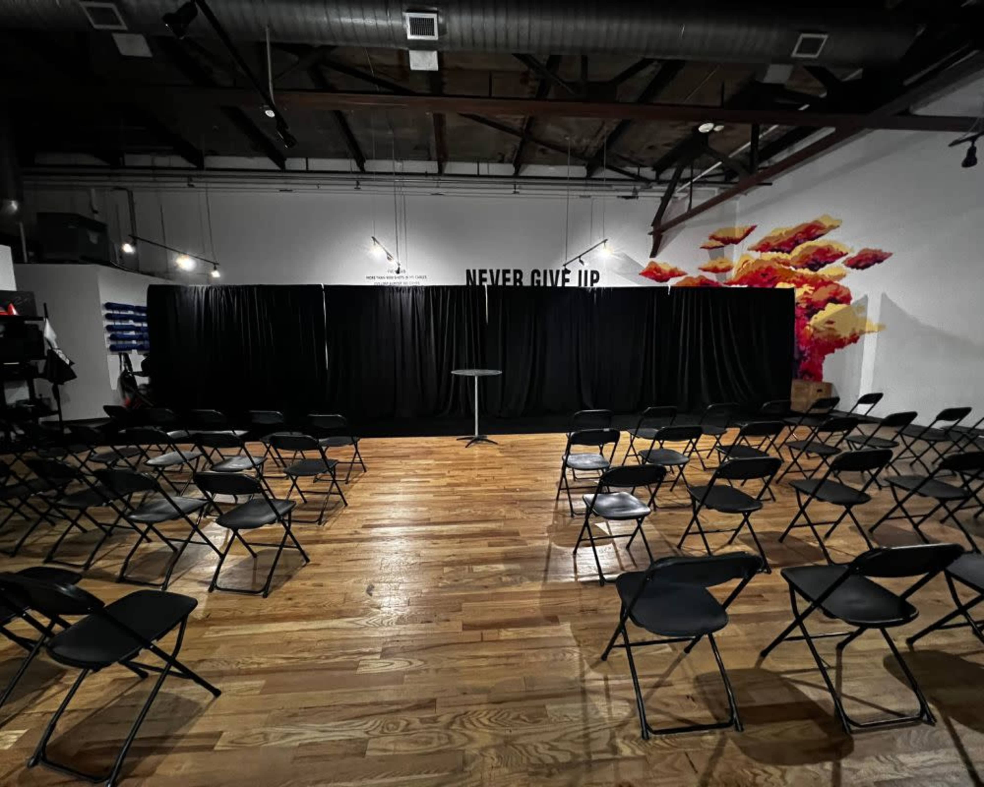 The scene shows a sparsely populated event space with rows of black chairs facing a small stage draped in black curtains, and a wall art featuring the words "NEVER GIVE UP."