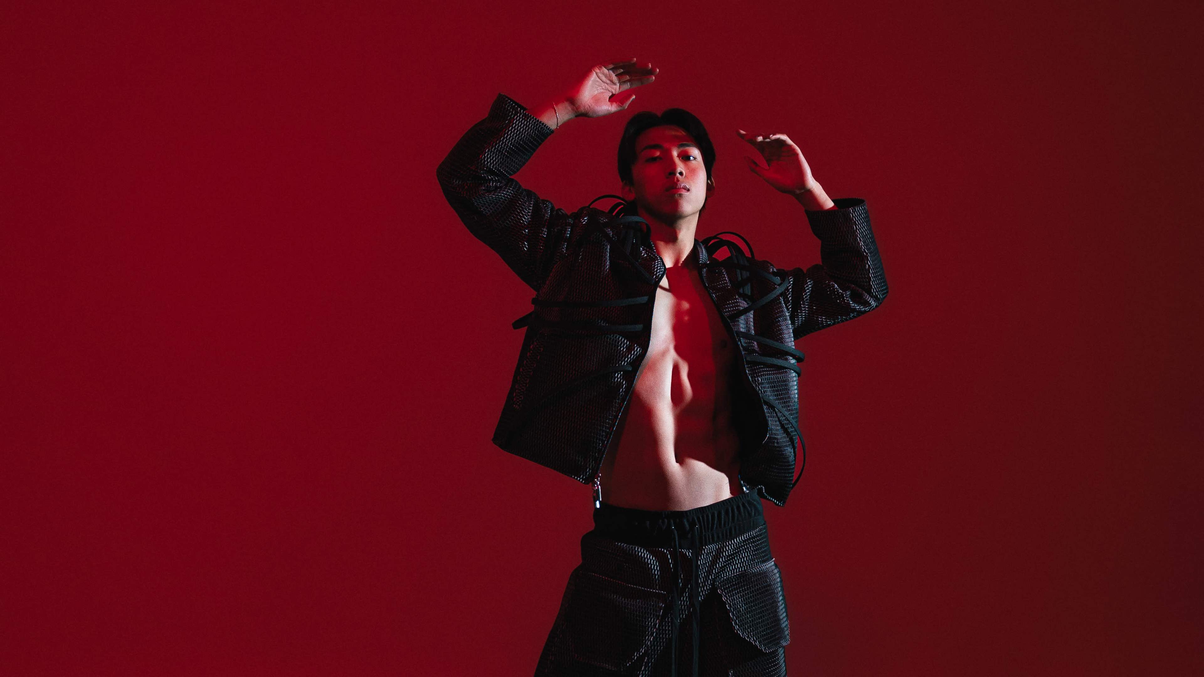A person poses with their arms raised against a solid red background, wearing a stylish black outfit.