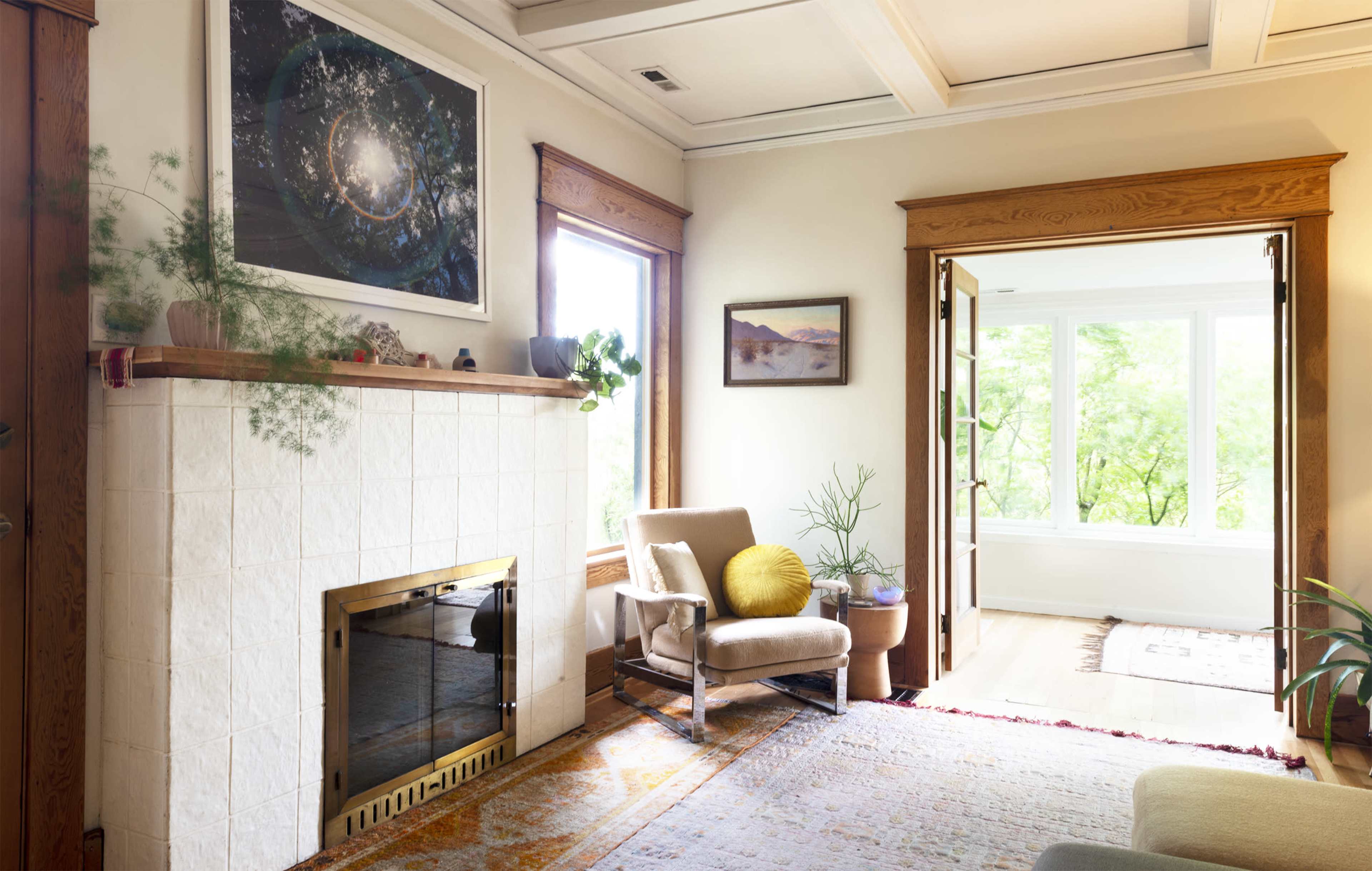 A bright living room featuring a white tiled fireplace, a modern chair with a yellow pillow, and large windows allowing natural light to fill the space.