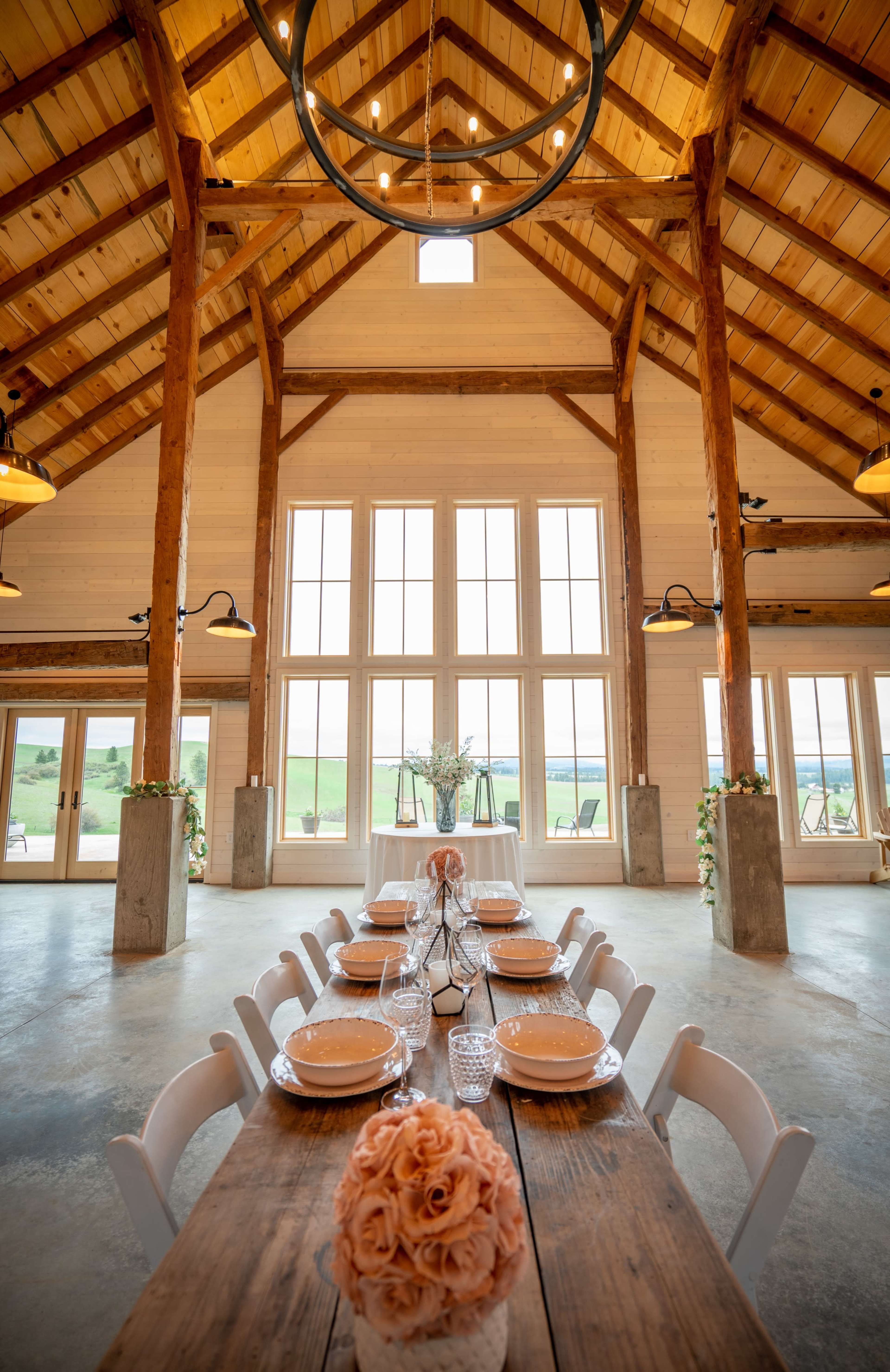A long wooden dining table is elegantly set with plates and glassware in a spacious room featuring high wooden beams and large windows.