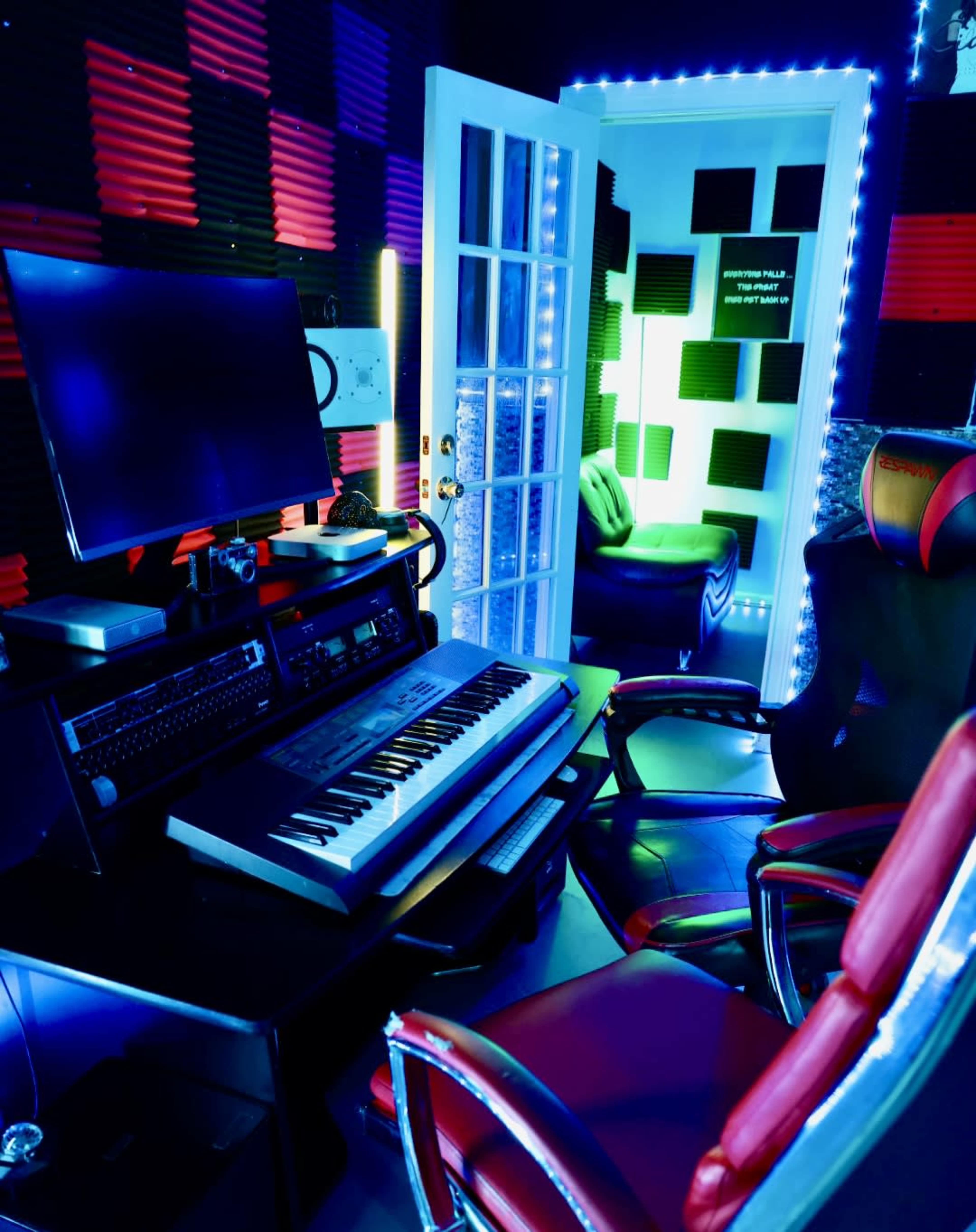 A music studio features a keyboard, computer monitor, and modern chairs with colorful lighting against acoustic foam walls.