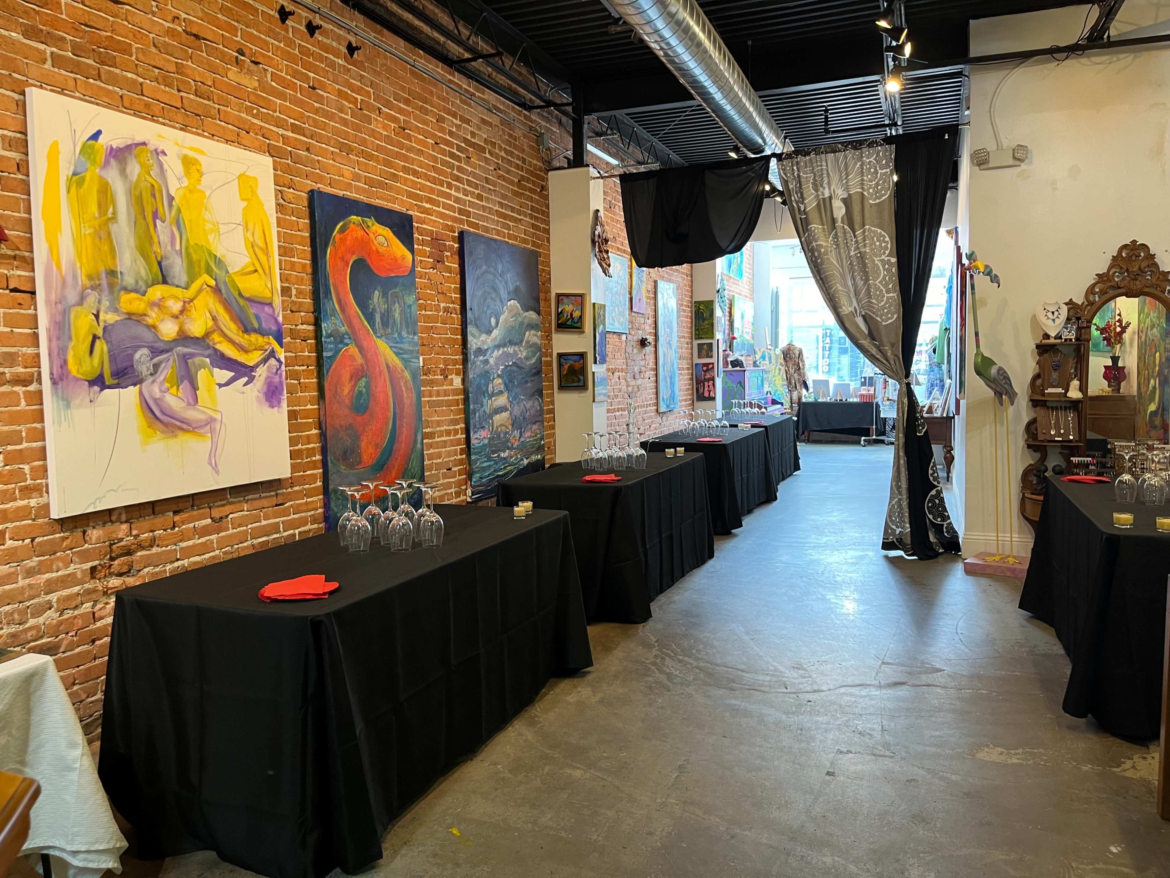 A gallery space featuring exposed brick walls, tables covered with black cloths, and various artworks displayed along the walls.