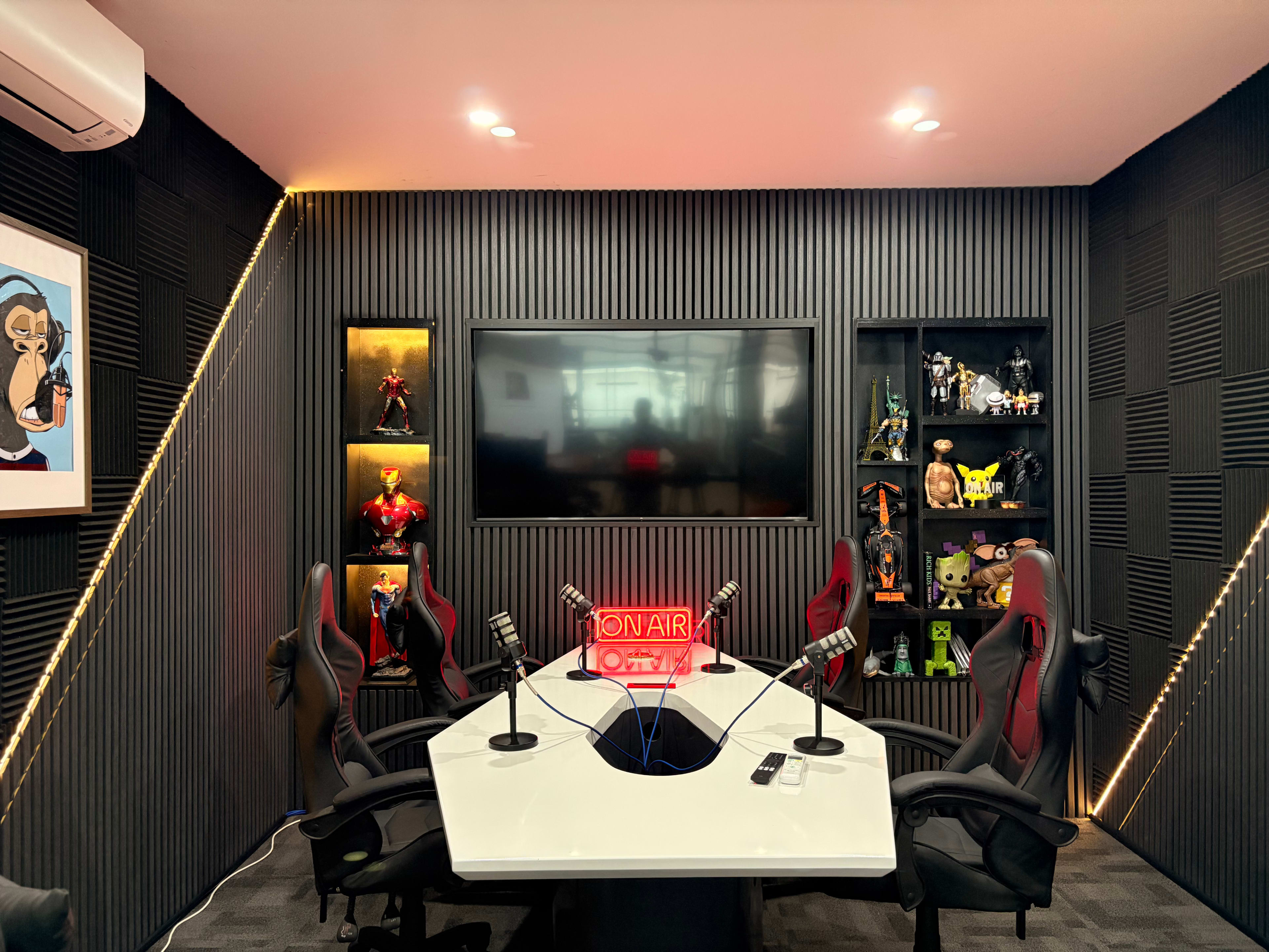 The image shows a modern podcast studio with a central white table, three microphones, black walls with vertical grooves, a large screen, and various figurines displayed on shelves.